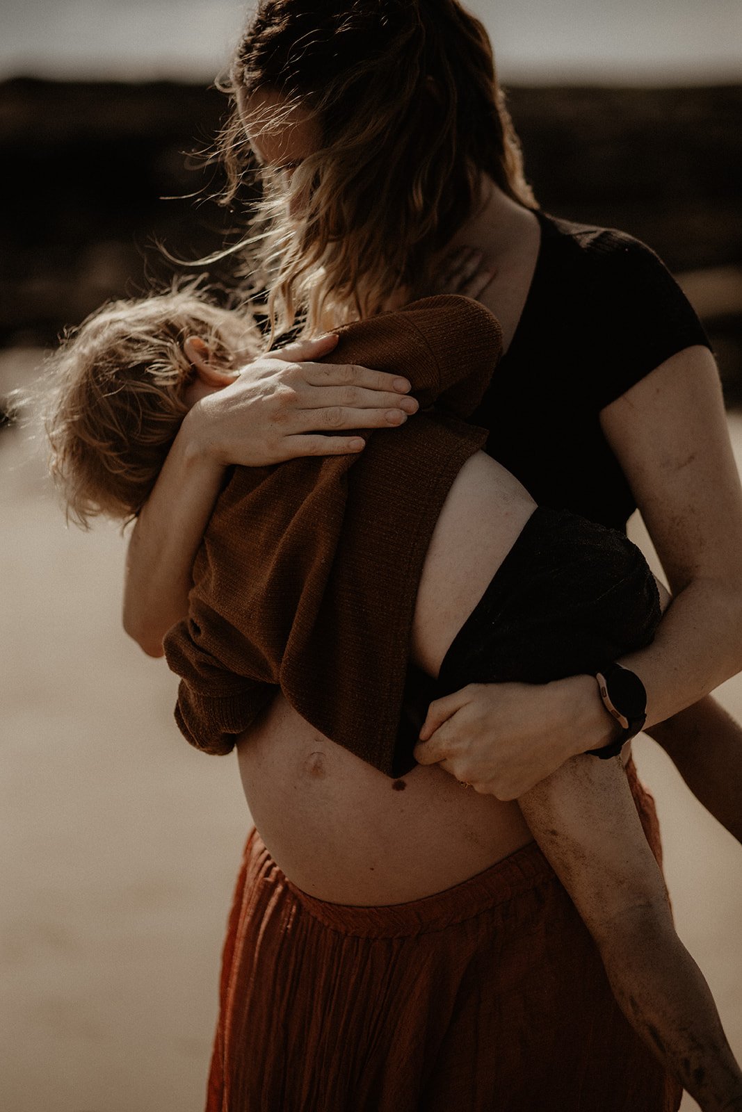 A woman holds a young child as they embrace on a sandy beach, with the woman wearing a black crop top and the child wrapped in a brown sweater.
