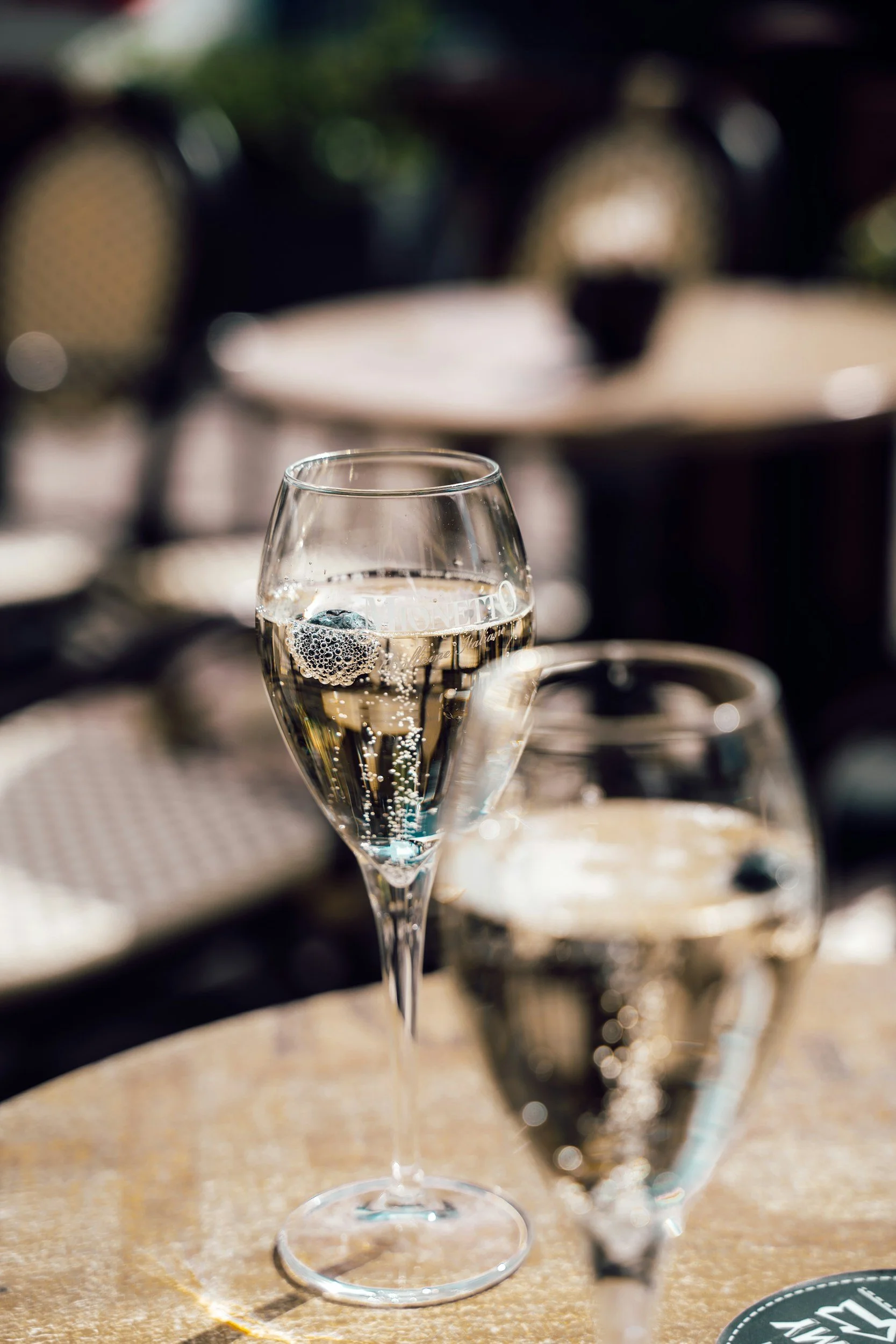 Two glasses of champagne on a table outside, with a blurred background of chairs and trees.