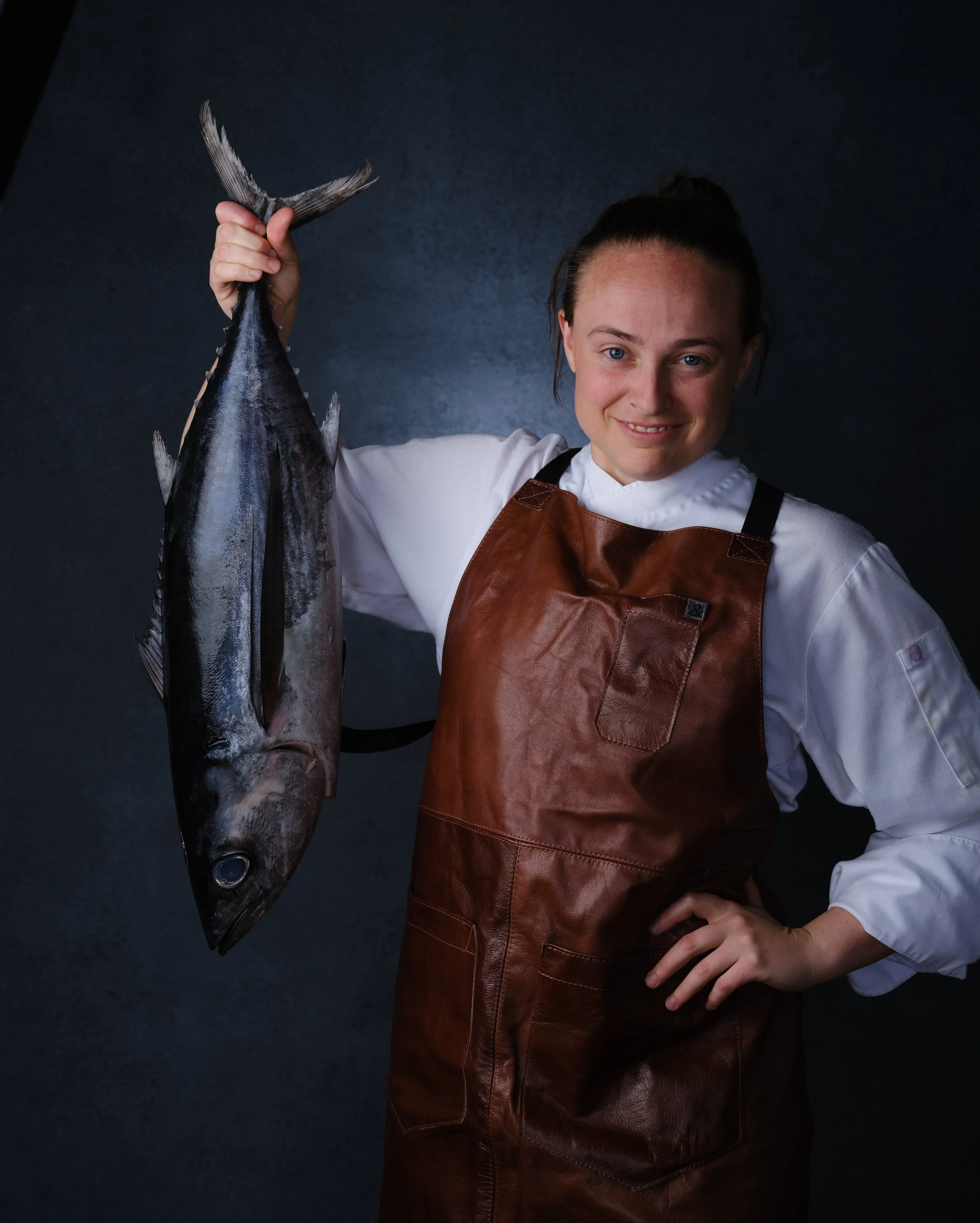 Young woman in chef's coat and apron holding a large fish against a dark background.