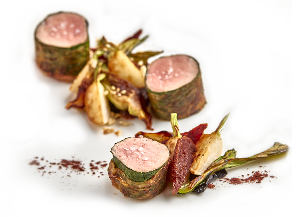 A gourmet dish featuring sliced, seared pink meat wrapped in a green vegetable, served with roasted vegetables and garnished with sauce and spices on a white plate.