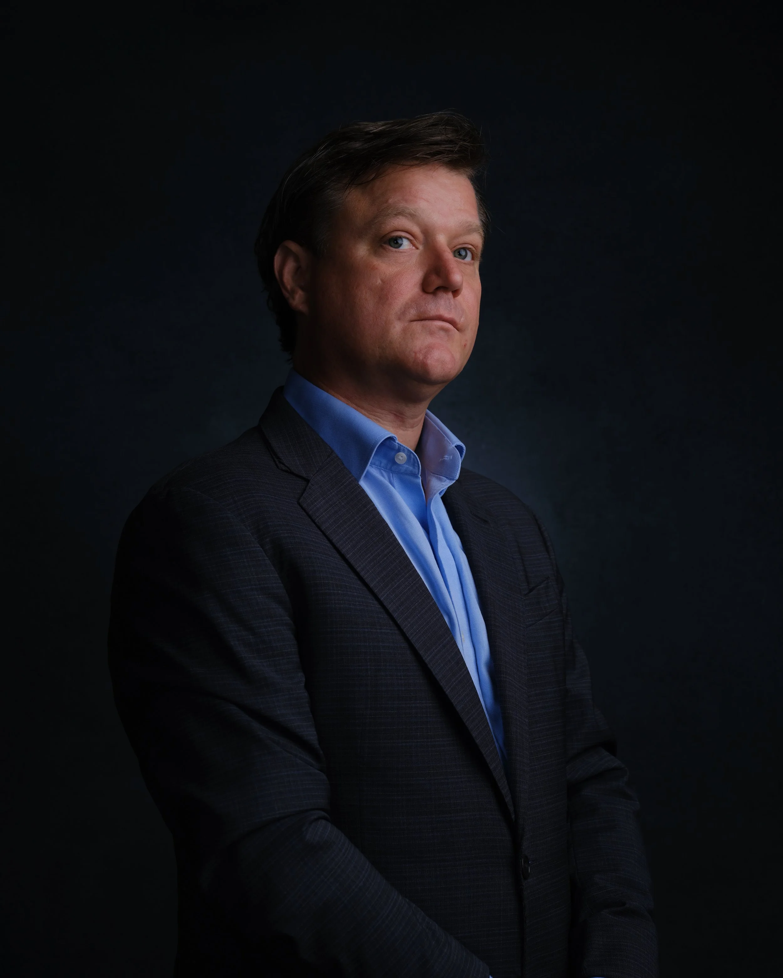 A man wearing a dark suit and light blue shirt poses against a black background.