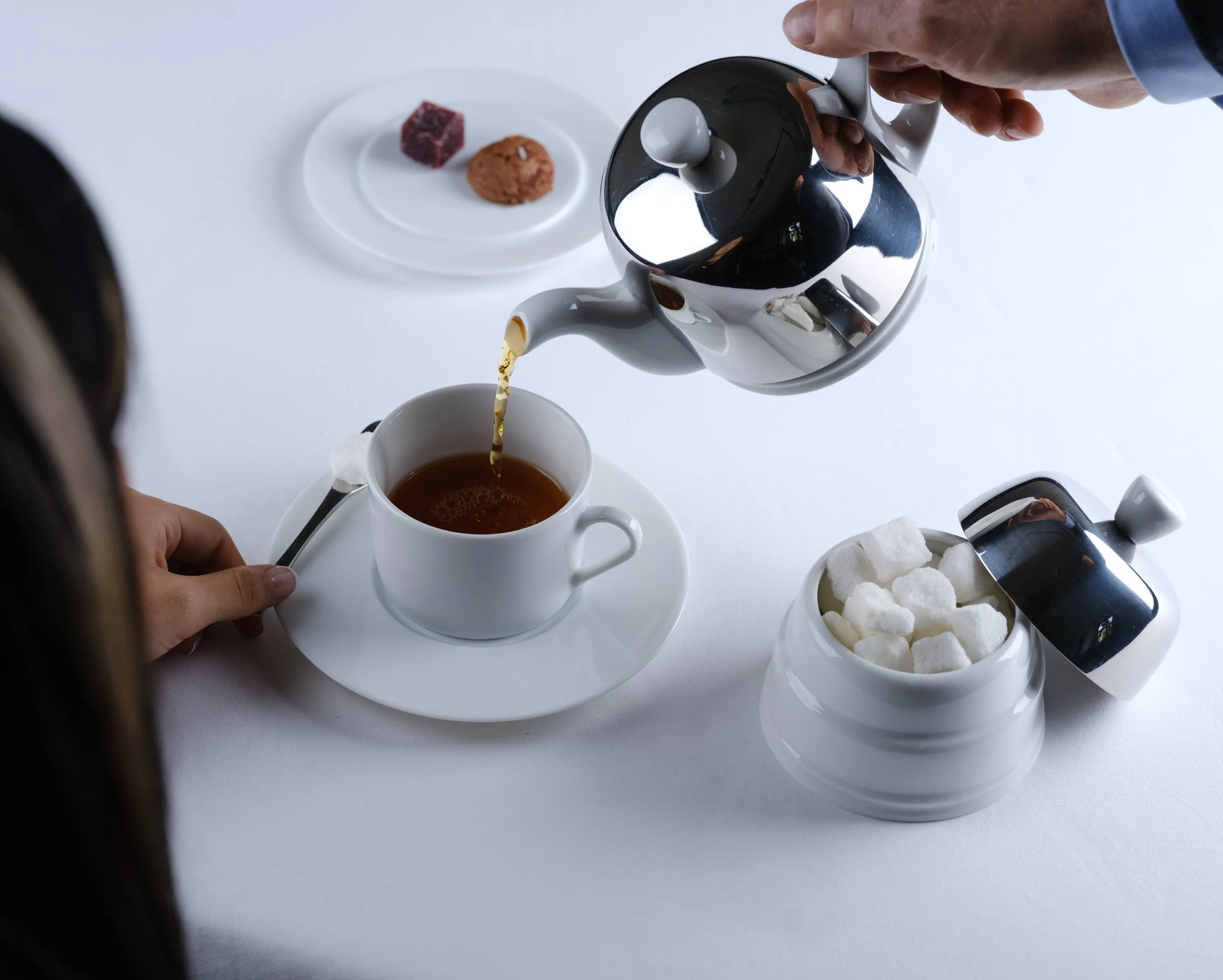 Person pouring tea from a silver teapot into a white teacup, with sugar cubes in a jar nearby and two types of cookies on a small plate in the background.