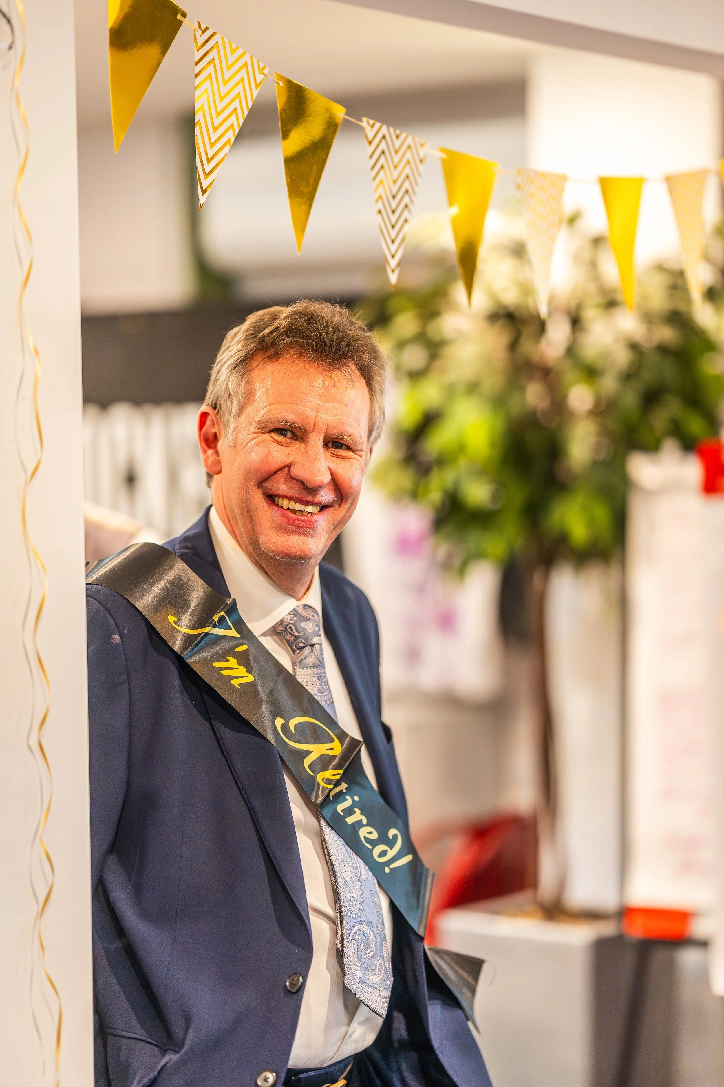 A man in a navy suit, light blue shirt, and orange tie stands next to a woman with blonde hair, wearing a black blazer over a patterned blouse, holding a colorful event flyer at a celebration of 15 years for Wolowise Bus for a Sport event, with a vibrant, multicolored backdrop.