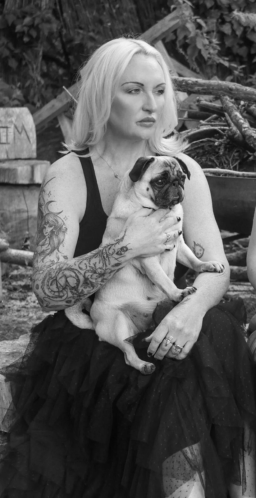A woman with tattoos on her arms, wearing a black top and skirt, sitting outdoors and holding a small pug dog.