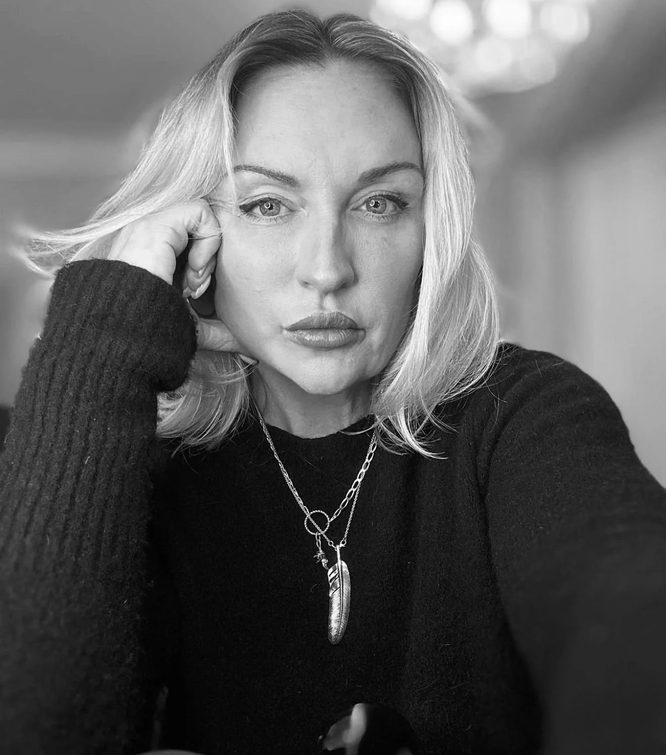 A woman with shoulder-length blonde hair, wearing a black sweater, resting her head on her hand, with two necklaces featuring pendants, in a black and white portrait.