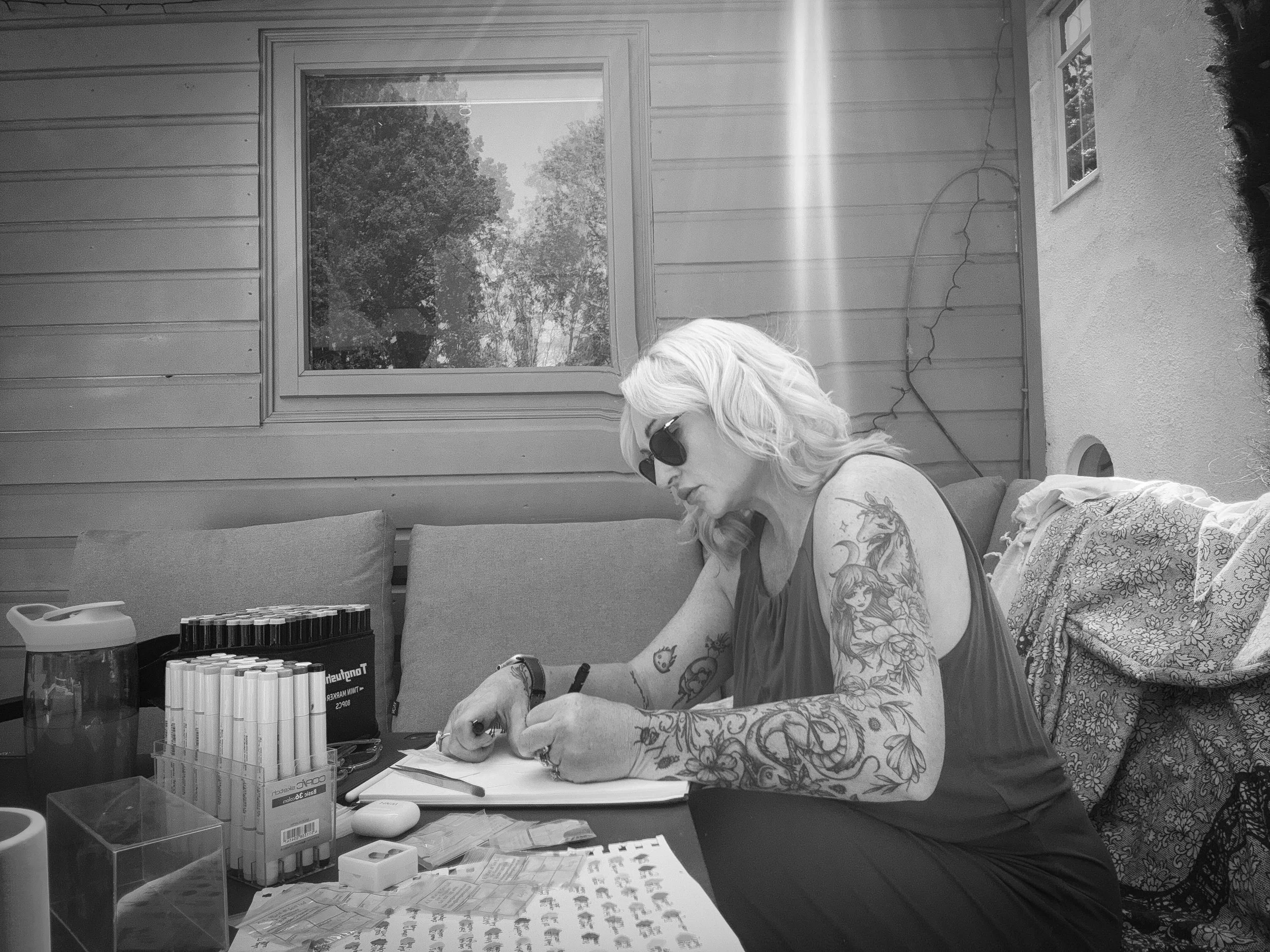 A woman with blonde hair, sunglasses, and tattoos on her arms, sitting at an outdoor table writing on a piece of paper, with various pens and markers, a water jug, and other supplies.