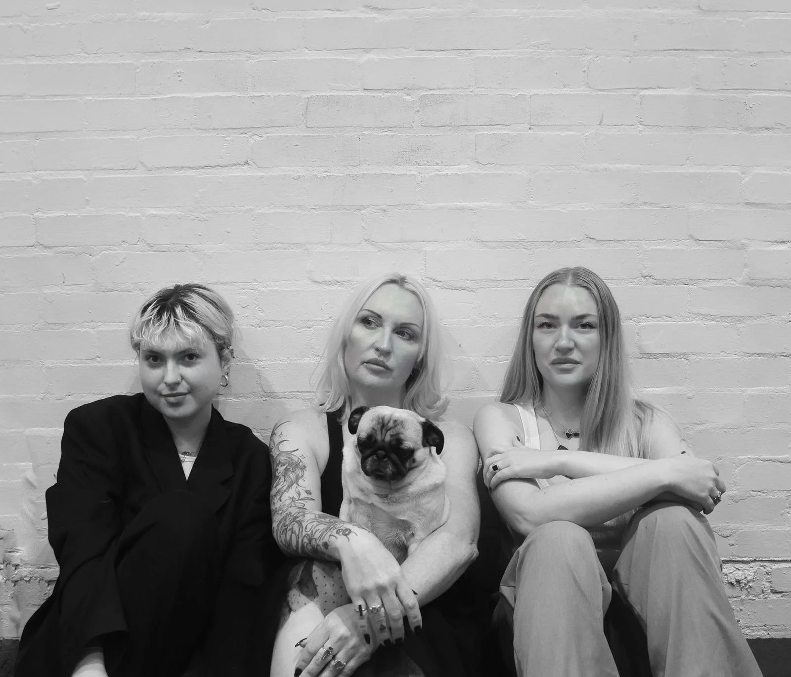 Three women sitting against a white brick wall, with one holding a pug dog while the others look at the camera with neutral expressions.