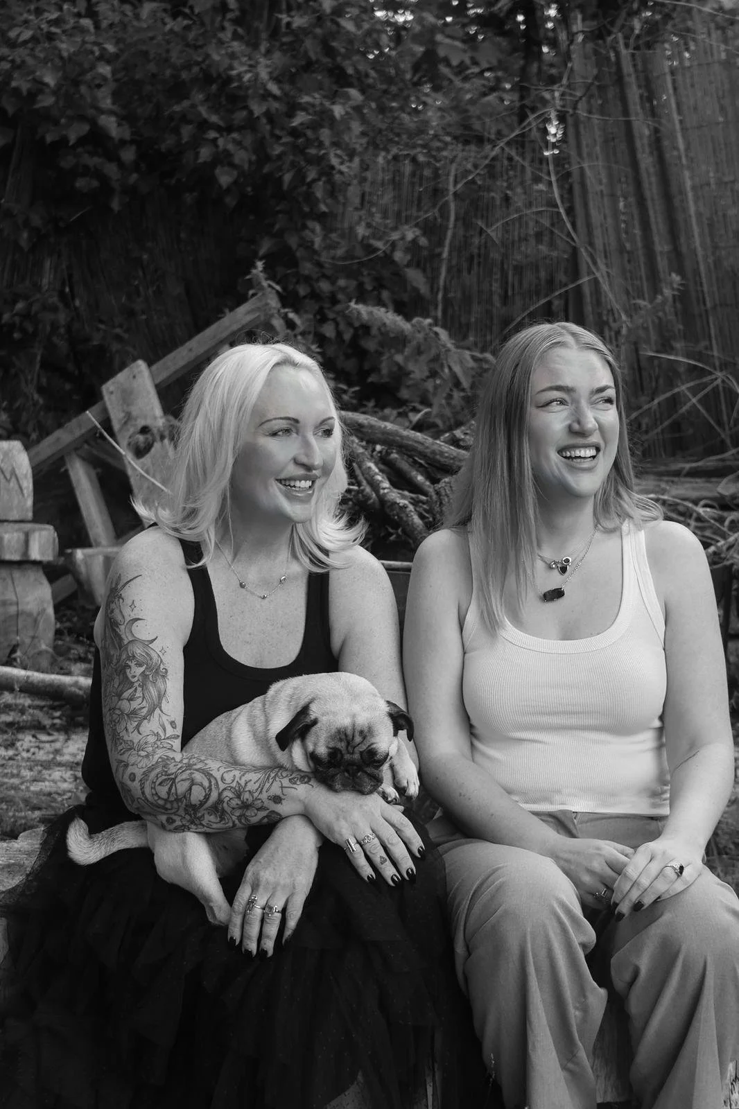 Two women sitting outdoors with a dog, smiling and looking in different directions, surrounded by trees.