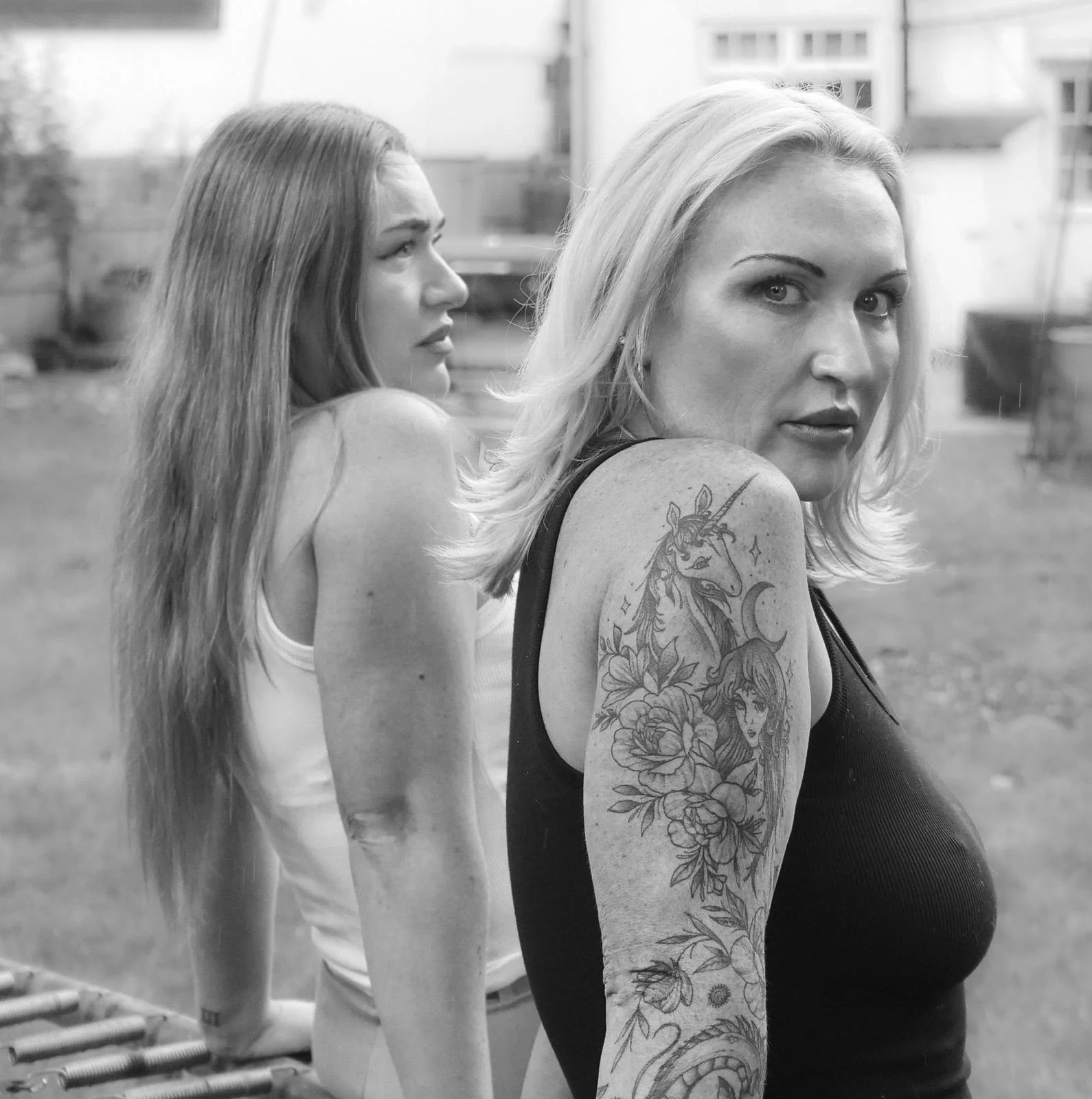 Two women standing outdoors in a black and white photo, one with long straight hair, the other with short wavy hair, the woman with short hair has tattoos on her arm, and is wearing a sleeveless top, while the woman with long hair is in a sleeveless