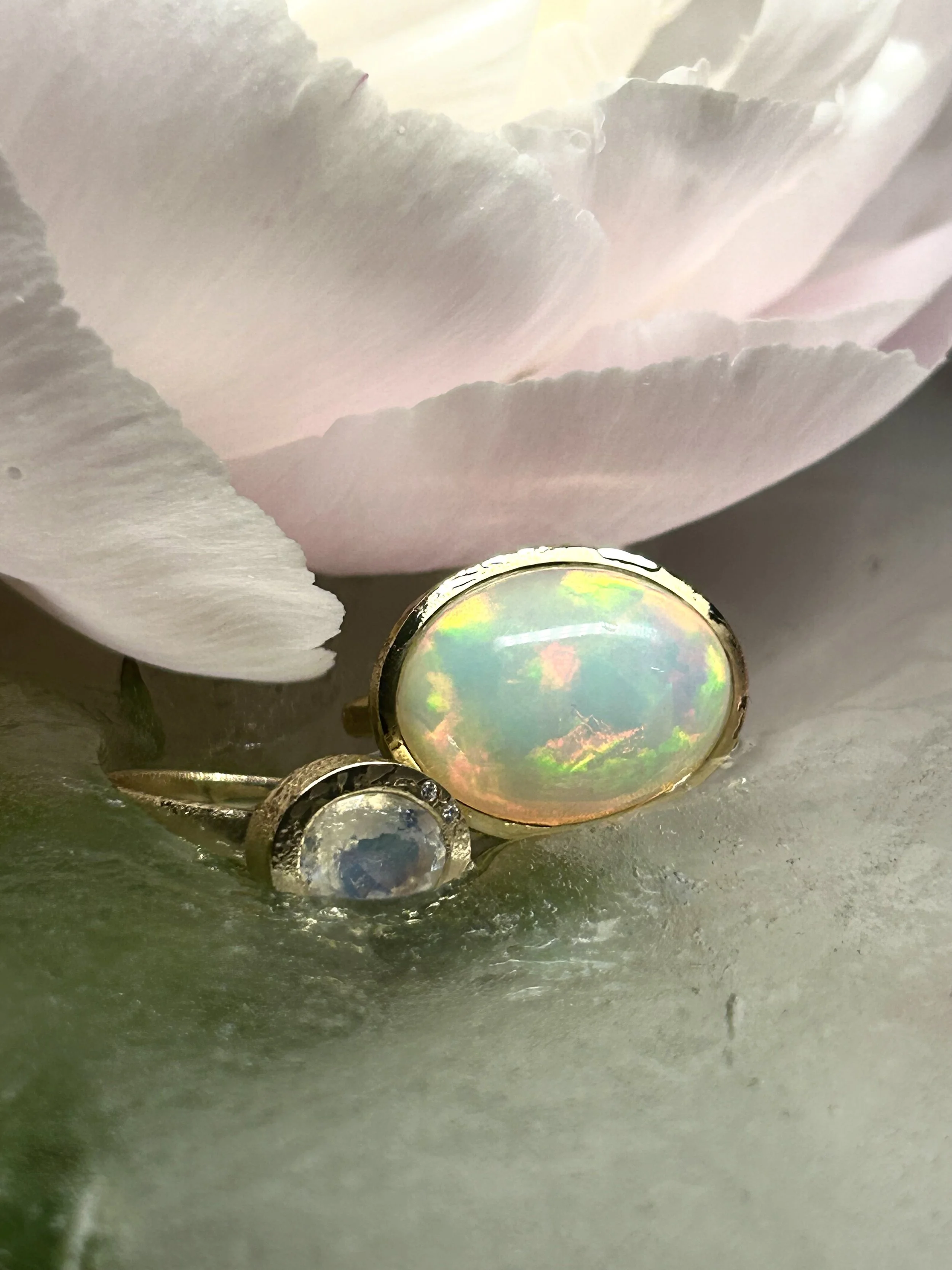 Two gemstone rings, one with an opal and the other with a clear stone surrounded by small diamonds, resting on a1 greenish surface next to a pink flower with soft petals.