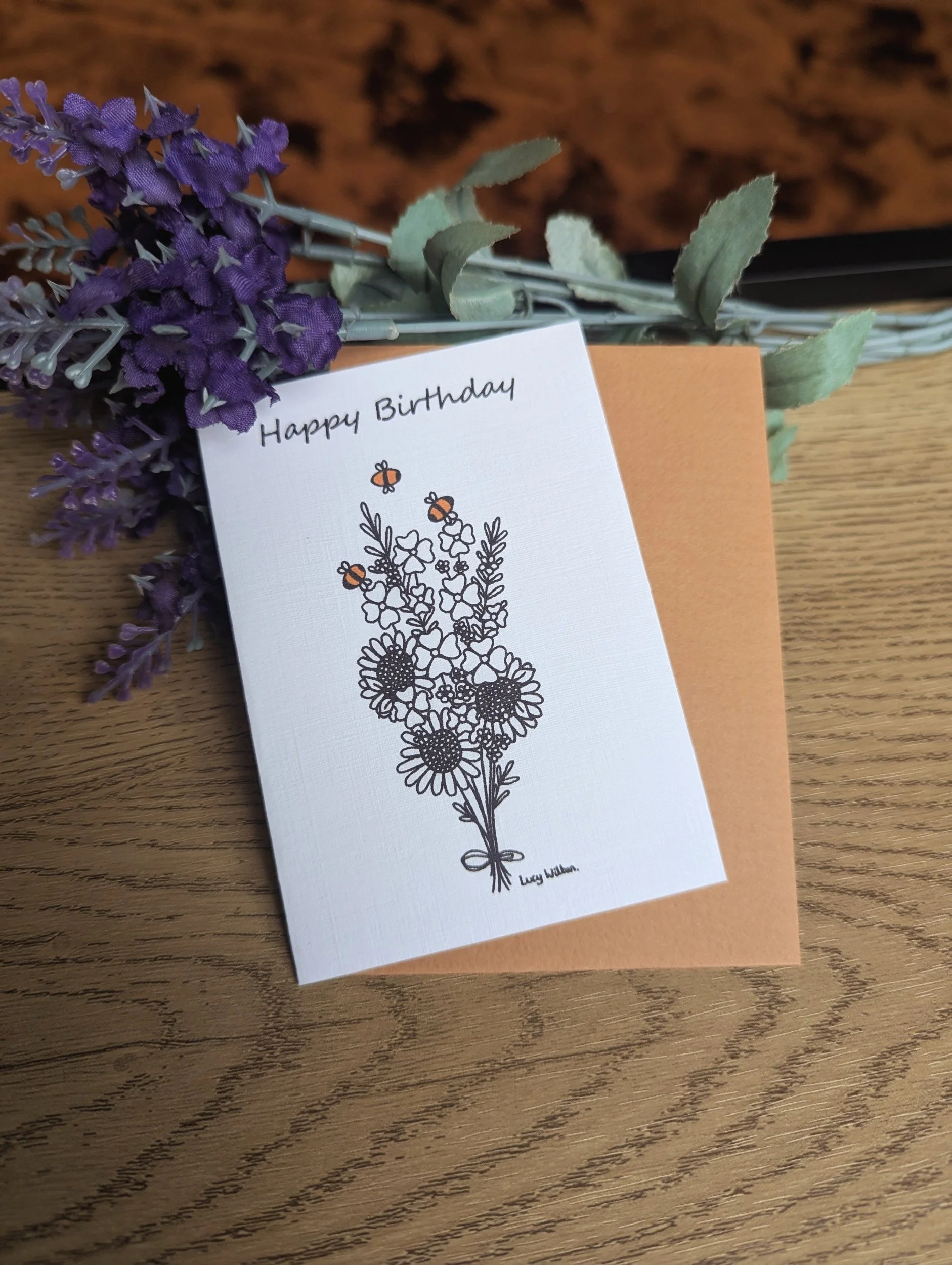 A purple lavender bouquet with green leaves and a greeting card that reads "Happy Birthday" with an illustration of a bouquet of flowers and bees, on a wooden surface, by Lucy Wilton Drawings
