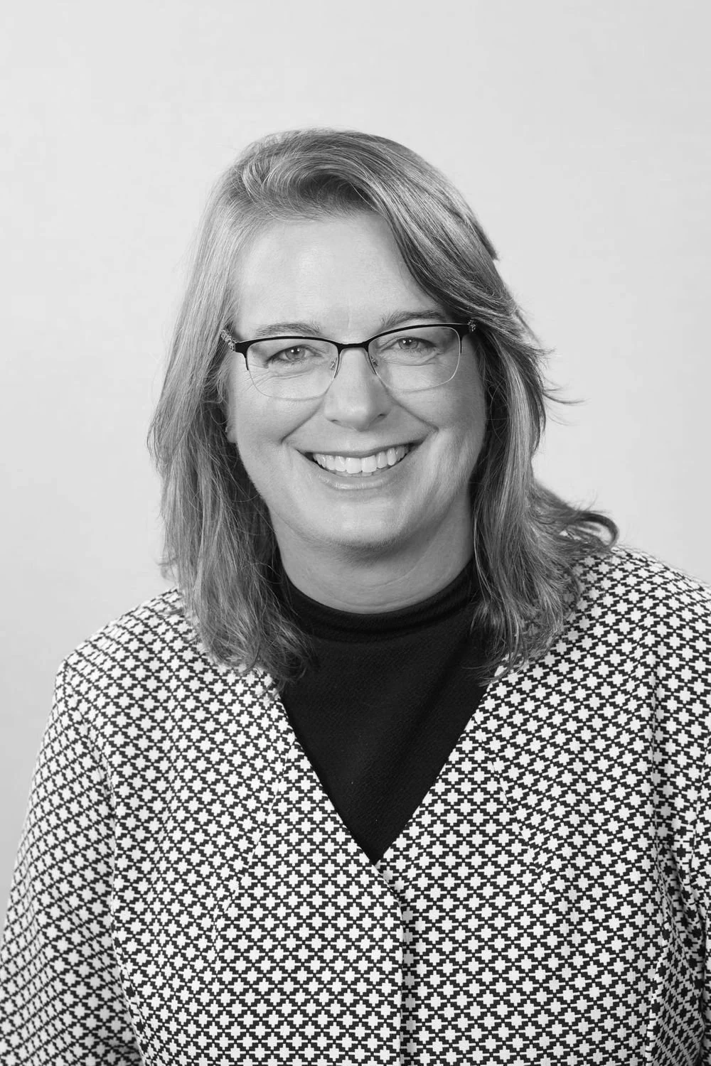 Black and white portrait of a woman with glasses, smiling, wearing a houndstooth blazer and a black turtleneck.