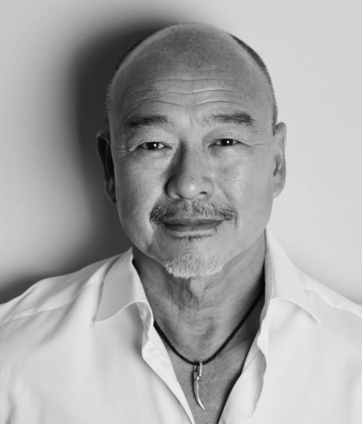 Black and white portrait of a smiling middle-aged Asian man with a shaved head, facial hair, wearing a white shirt and a black necklace with a pendant.
