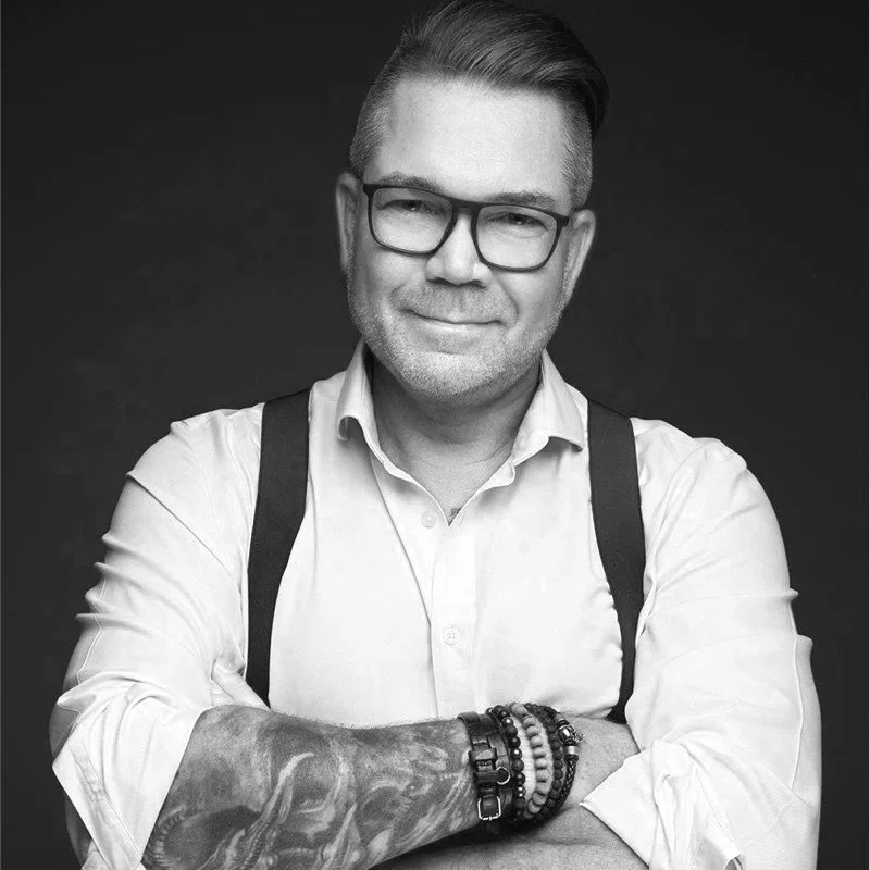 Nick Shakelton Jones - Black and white portrait of a man wearing glasses, a light-colored button-up shirt, suspenders, and multiple bracelets on his left wrist, smiling slightly with arms crossed against a dark background.