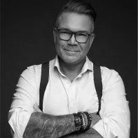 Portrait of a man with glasses, wearing a white shirt, with his arms crossed, and a tattoo on his arm, in black and white.