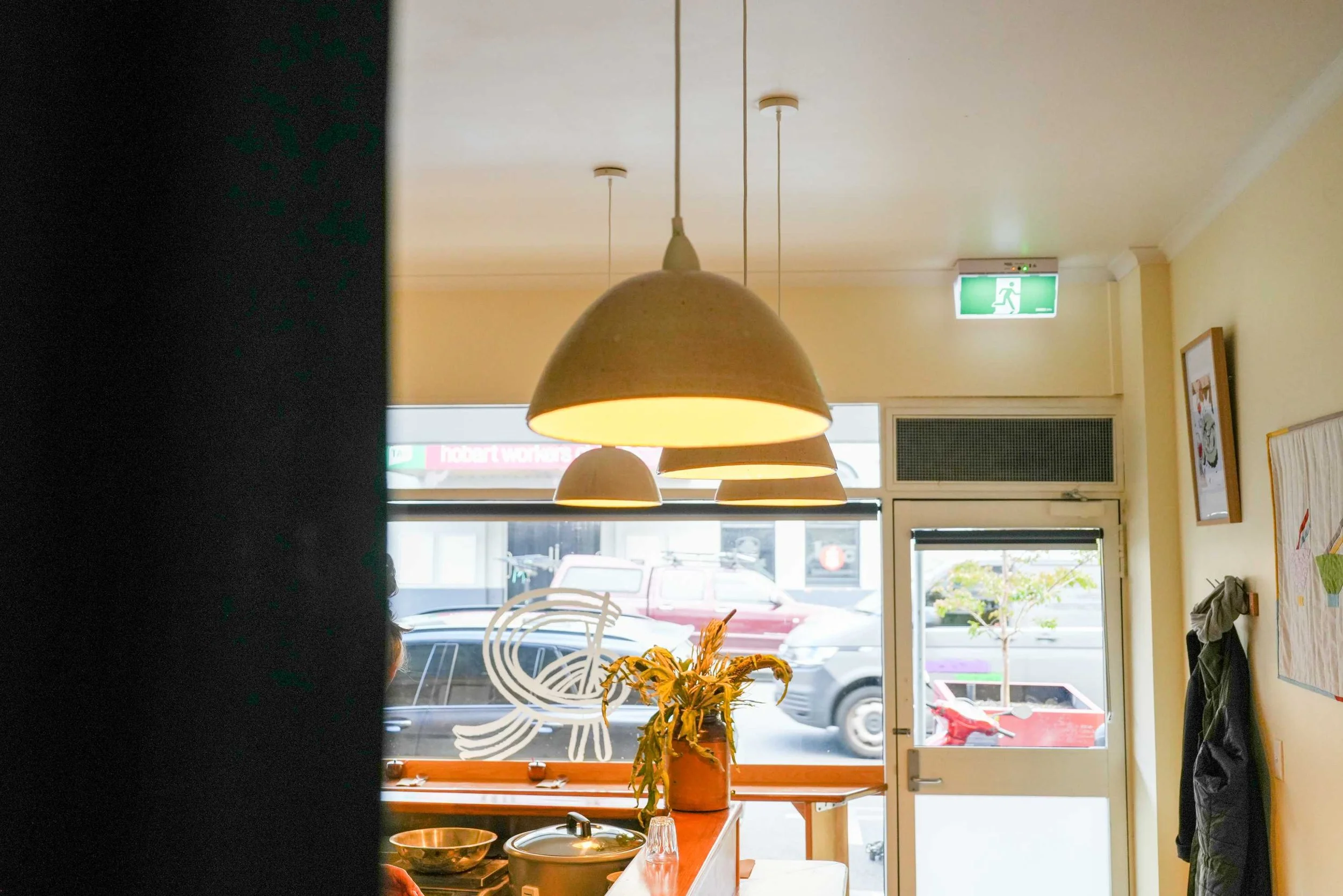 Interior of a cafe with hanging lamps, a potted plant on a counter, and a view of parked cars outside through large windows. There is an emergency exit sign on the ceiling.