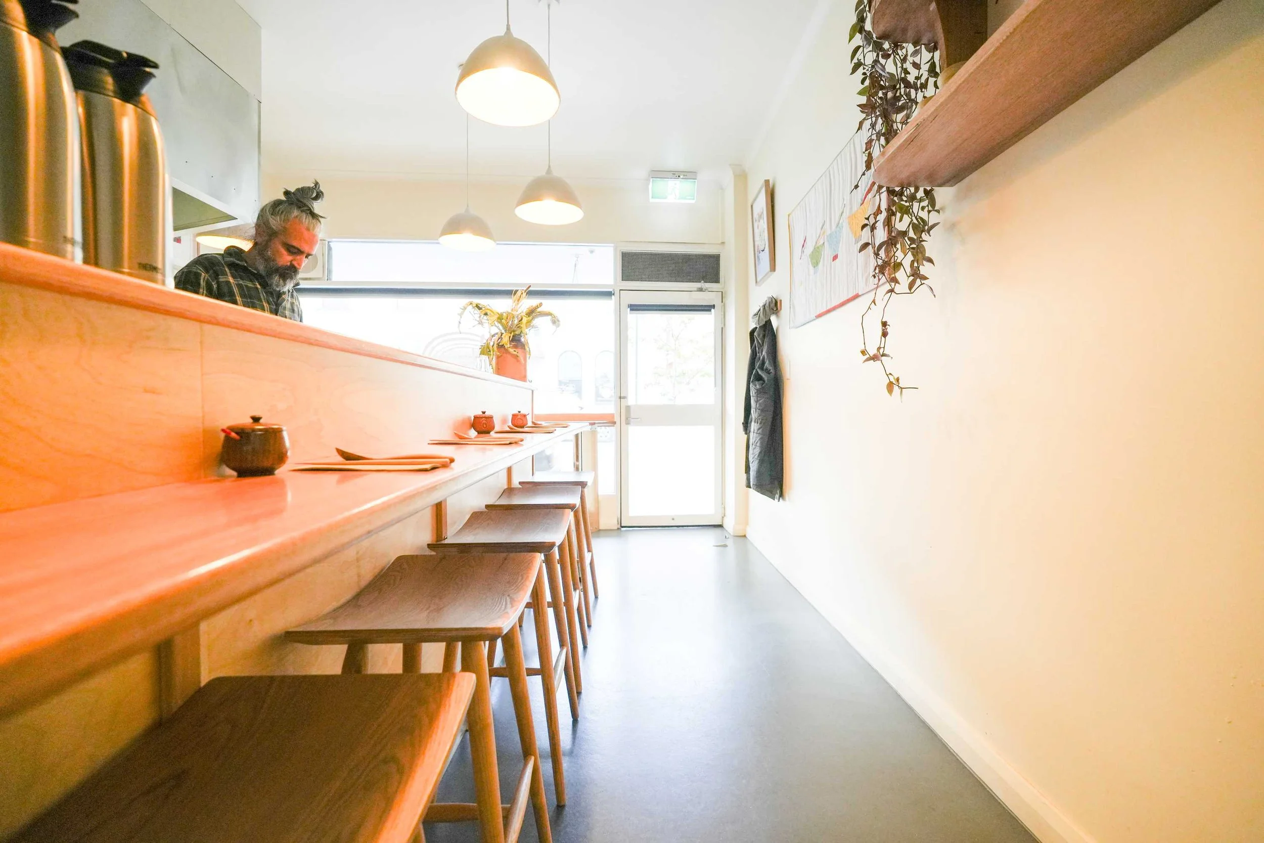 A man with long hair and a beard sitting inside a small, minimalist restaurant or café. The room features a wooden counter with bar stools, hanging pendant lights, a wall decoration, and a potted plant. There is a door at the back with a glass window, letting in natural light.