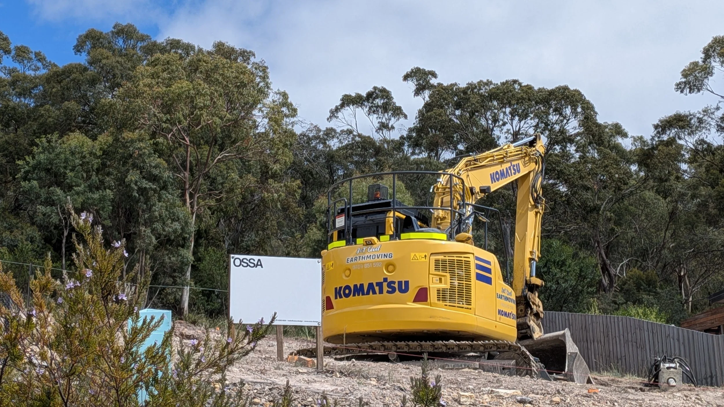 A yellow Komatsu excavator labeled "JET EARTH EARTHMOVING" and "KOMATSU" is on a construction site surrounded by trees. There is a white sign with black letters that reads "OSSA." The excavator is situated on a concrete slab with rocks and construction debris around.