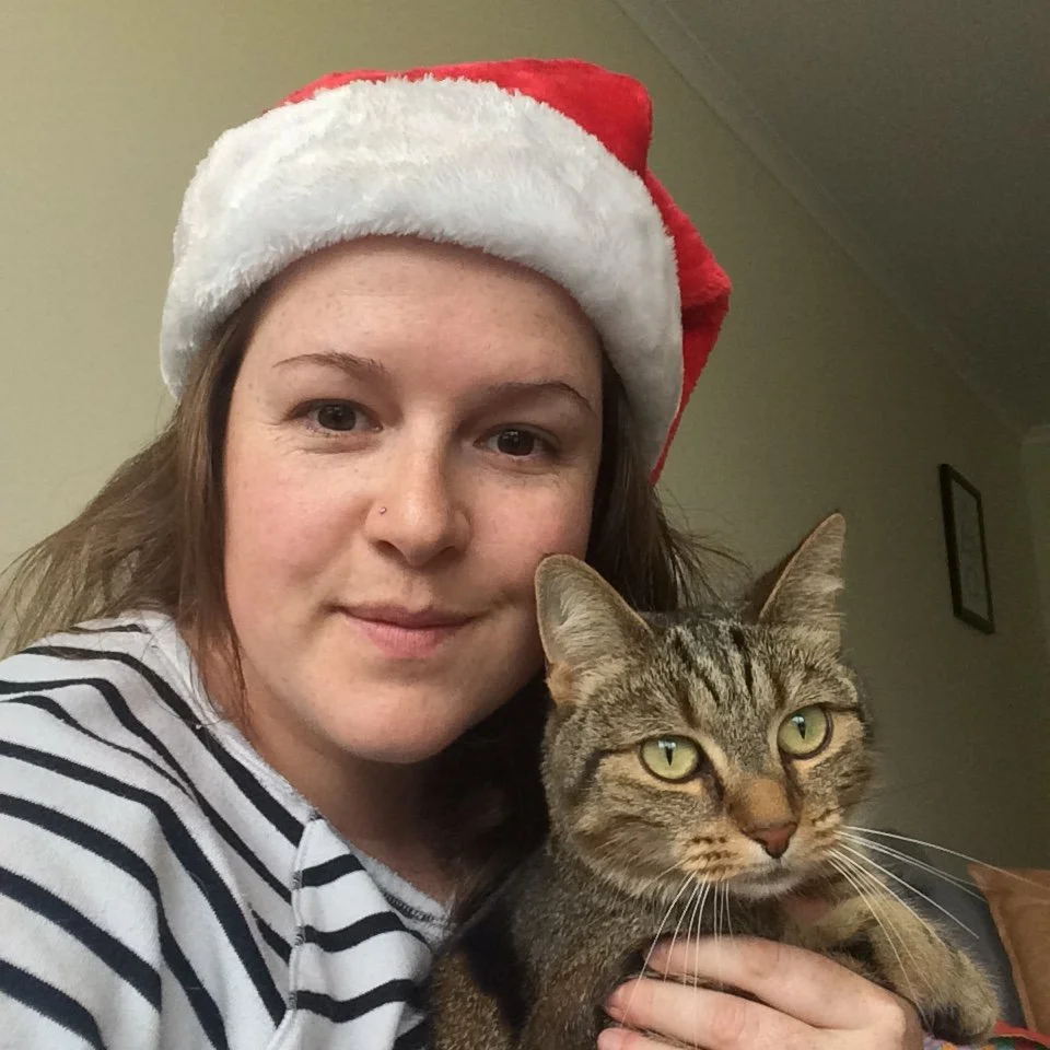 A woman wearing a Santa hat and a black and white striped shirt holding a tabby cat with green eyes.