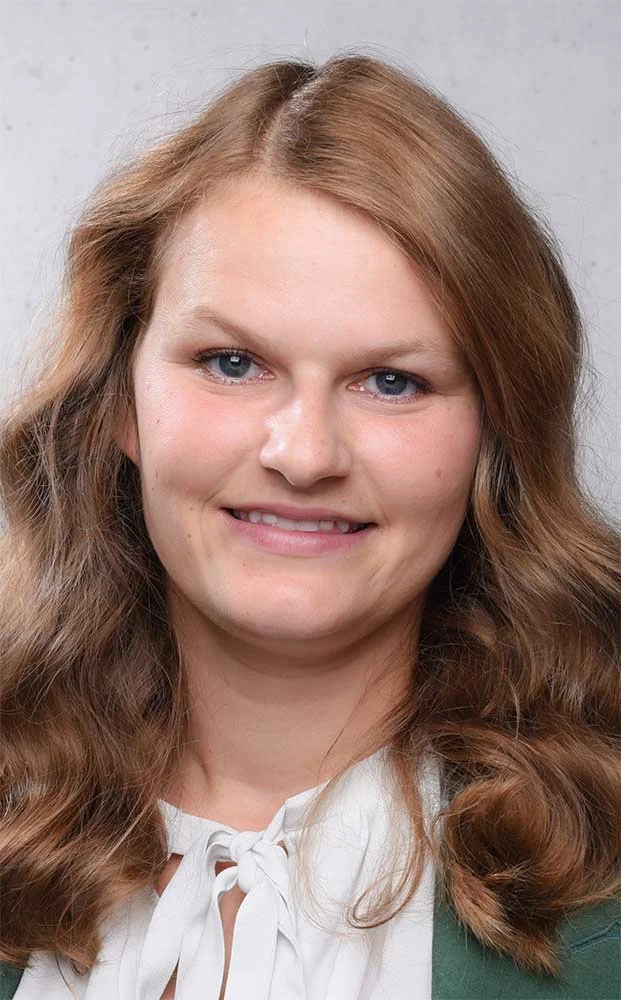 Close-up of a smiling young woman with long brown, wavy hair, blue eyes and fair skin, wearing a white blouse with a bow, in front of a light background.