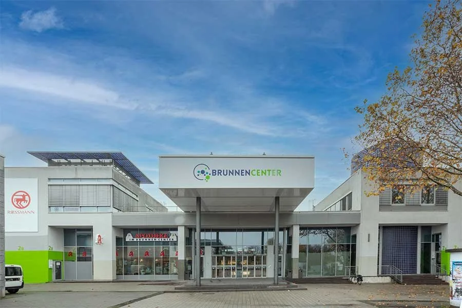 The image shows the exterior of a modern building with the sign 'BrunnenCenter' and other shops such as a pharmacy and Rossmann. It is sunny and there is a tree on the right side.