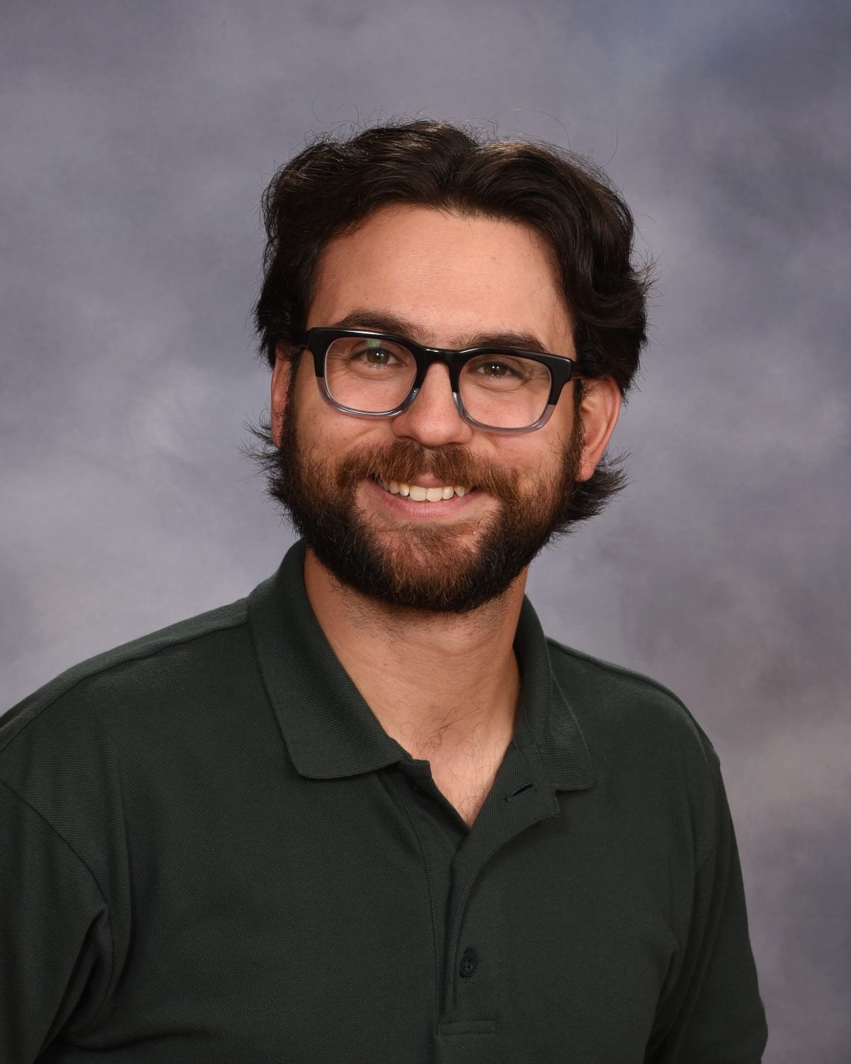 Portrait of a smiling man with glasses, dark hair, and beard, wearing a dark green polo shirt, against a gray background.