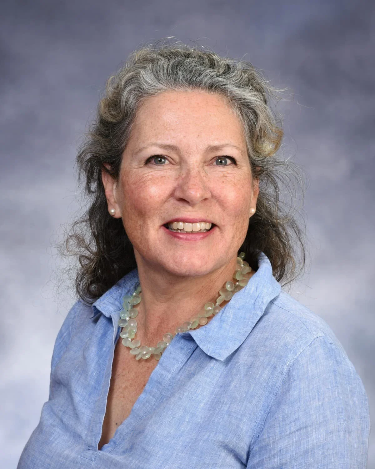 A portrait of a smiling middle-aged woman with curly gray hair, wearing a light blue blouse, pearl earrings, and a pearl necklace, posed against a neutral gray background.