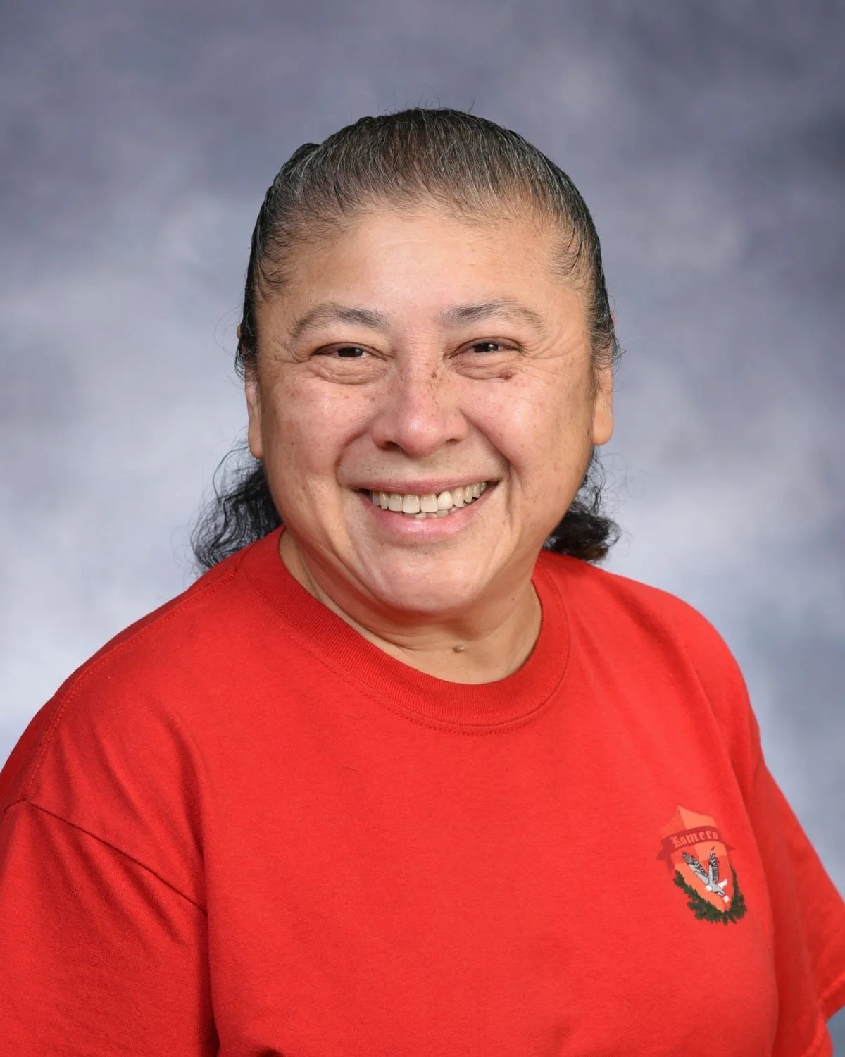 A woman smiling at the camera wearing a red shirt with a small logo on the chest. Background is a soft, cloudy gray.