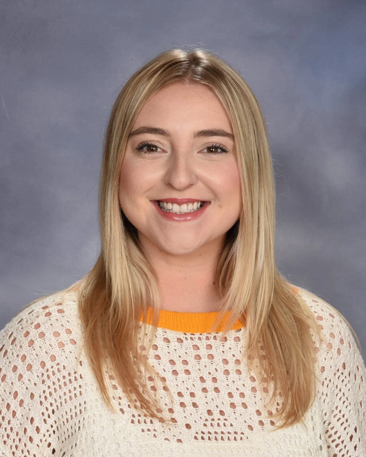 A woman with long blonde hair smiling at the camera, wearing a cream-colored knitted sweater with orange accents, against a gray background.