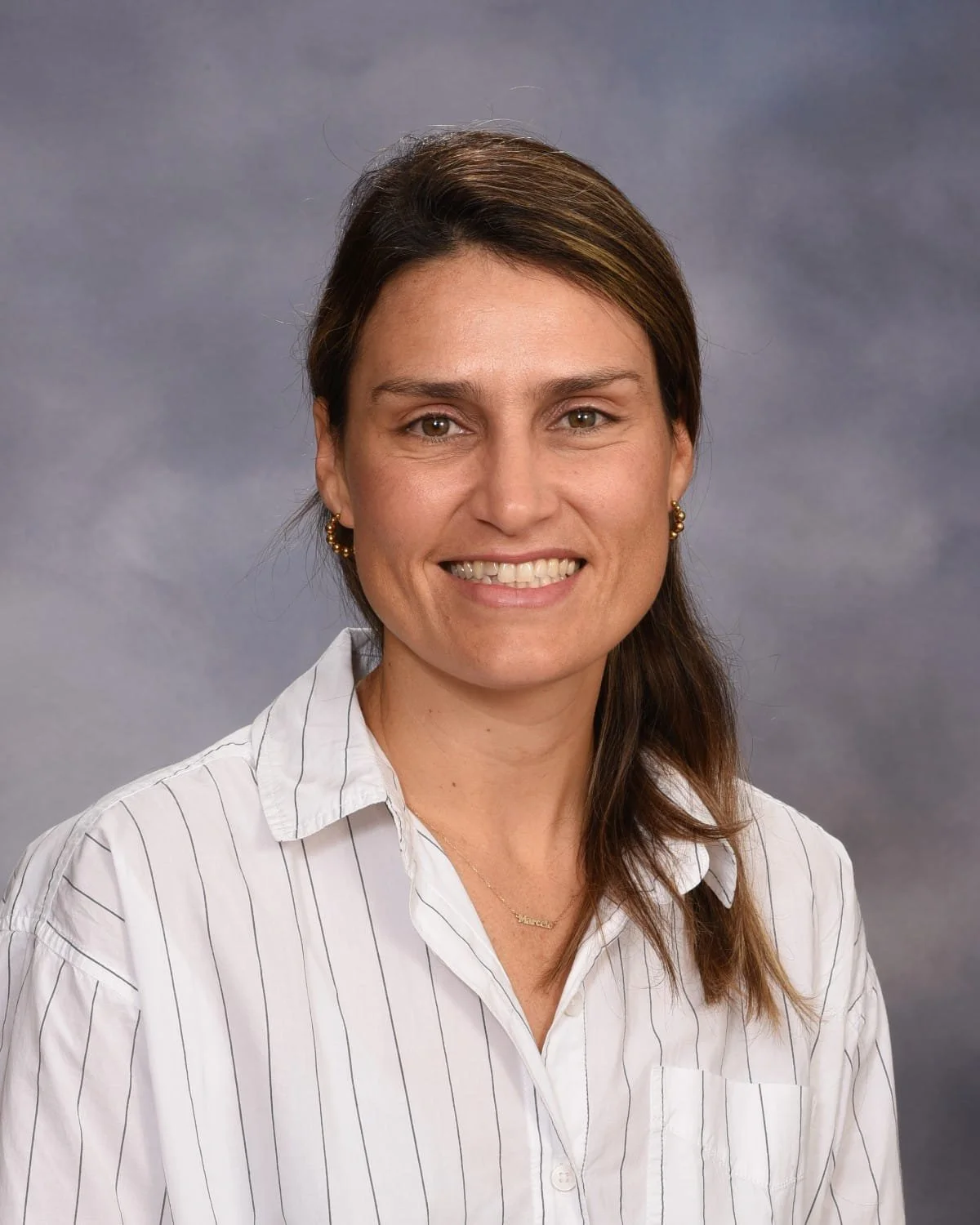 A woman with shoulder-length brown hair, wearing a white striped shirt, smiling against a gray background.