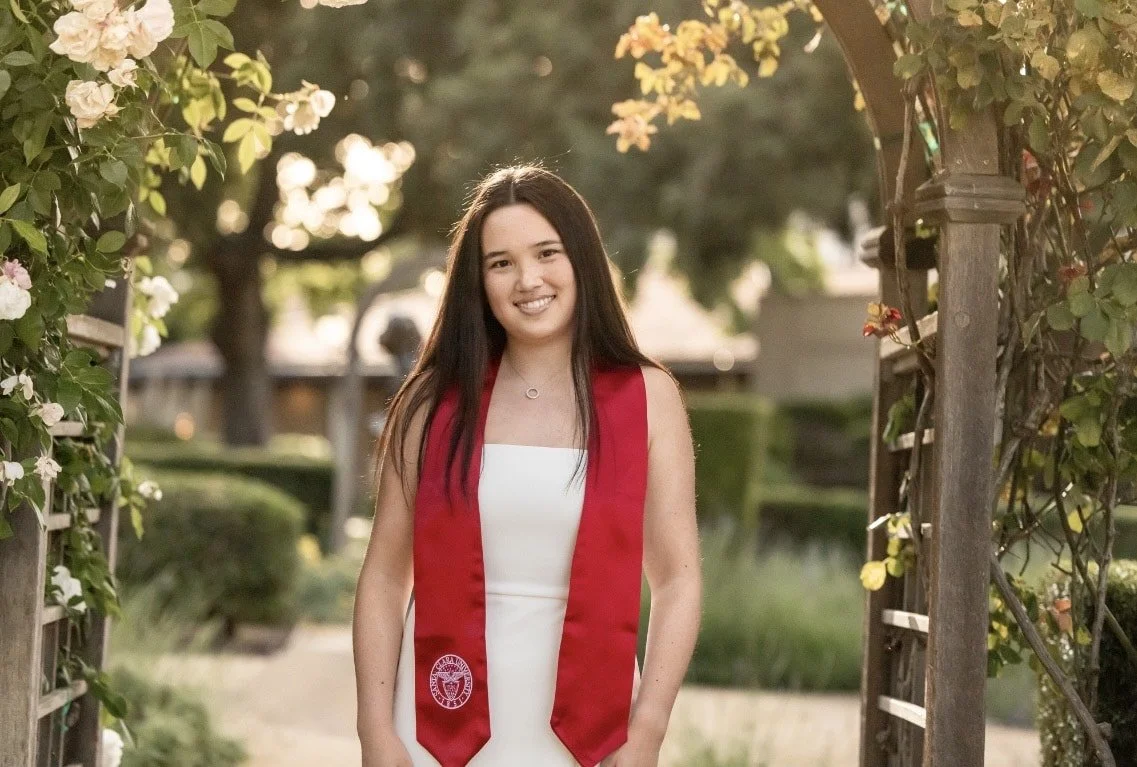 A young woman in a white dress and red graduation stole standing outdoors on a garden pathway, smiling with dark hair, surrounded by flowering plants and an arbor in the background.