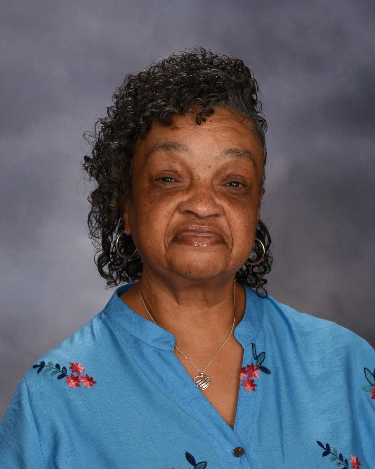 Portrait of a woman with dark, curly hair, wearing a light blue blouse with floral embroidery, hoop earrings, and a silver necklace with a circular pendant, against a dark grey background.