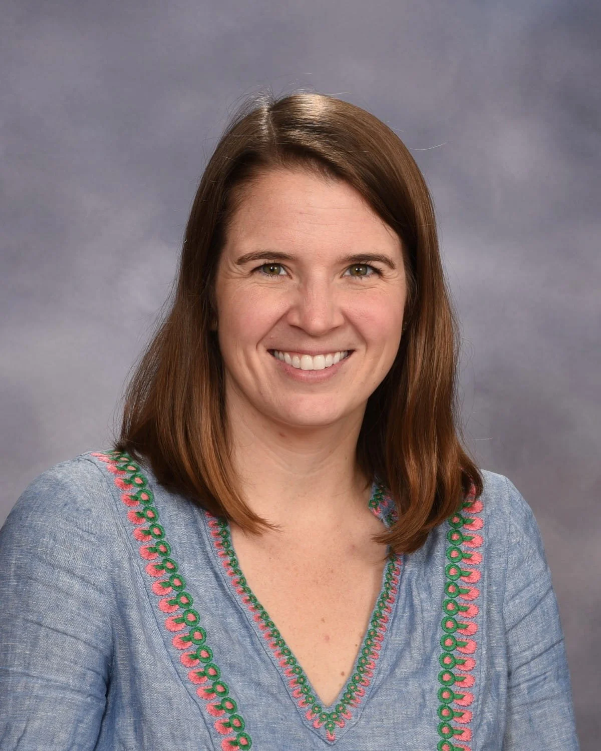 Portrait of a woman with shoulder-length brown hair wearing a blue top with embroidered pink and green details, smiling against a gray background.
