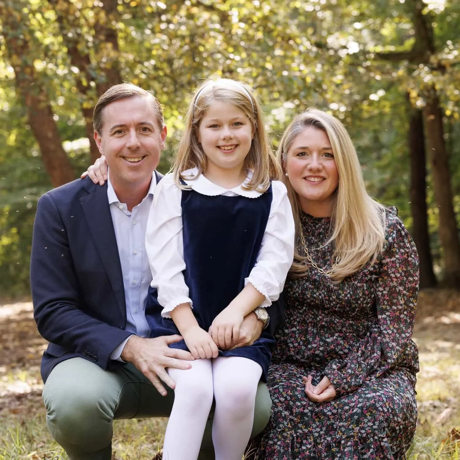 A smiling family of three sitting together outdoors in a forested area, with autumn leaves and sunlight filtering through trees.