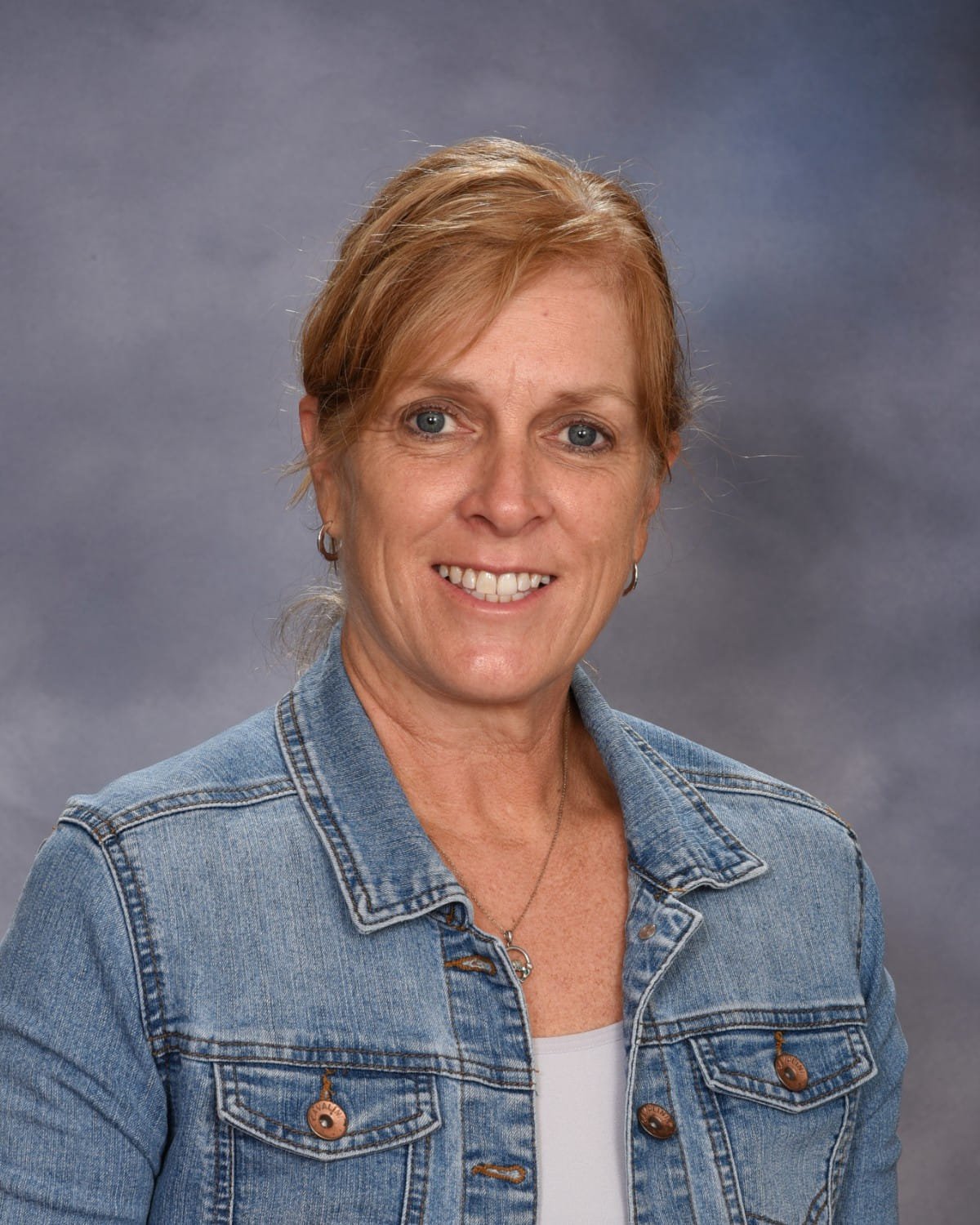 A woman with short red hair wearing a denim jacket and white top, smiling at the camera against a neutral gray background.