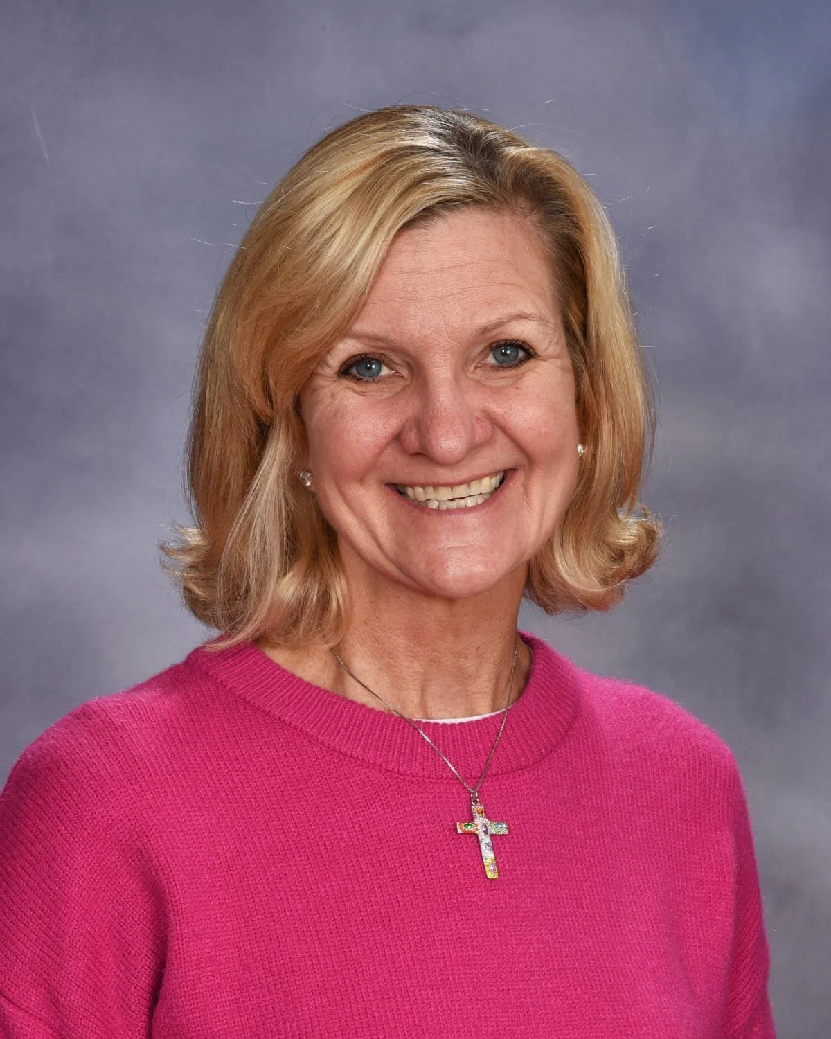 A woman with shoulder-length blonde hair, blue eyes, and earrings, wearing a pink sweater and a cross necklace, smiling against a gray background.
