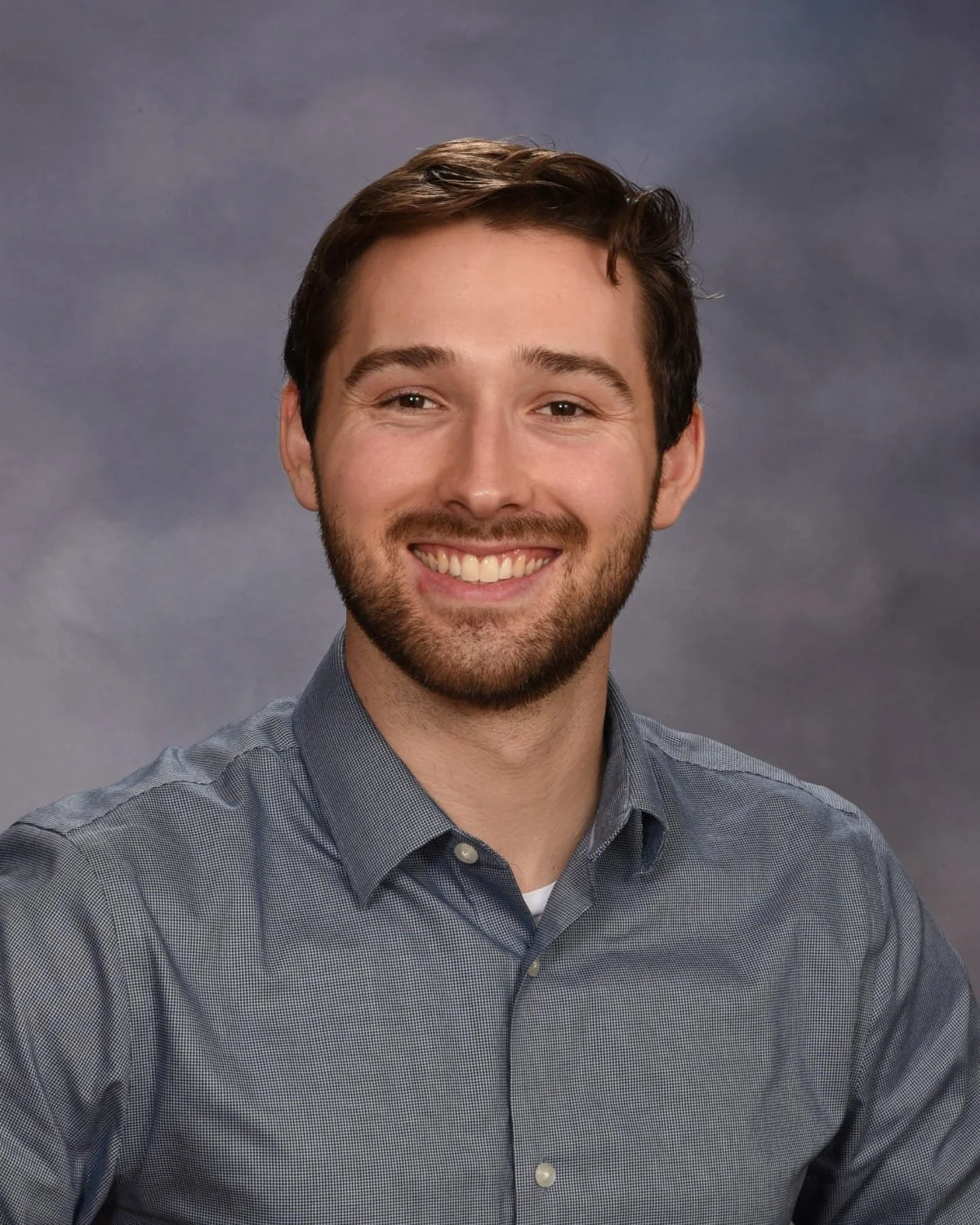 A man with brown hair and a beard, smiling, wearing a gray button-up shirt against a gray background.