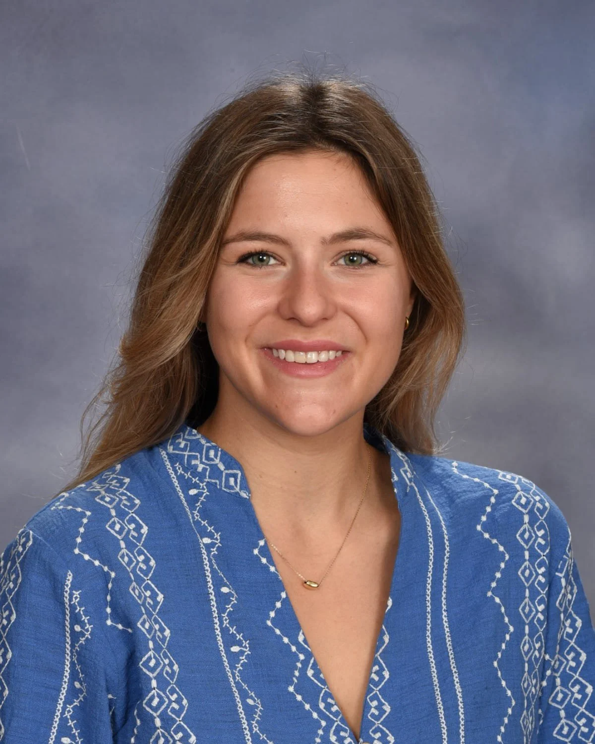 Headshot of a young woman with shoulder-length light brown hair, wearing a blue embroidered blouse and a gold necklace against a gray background.