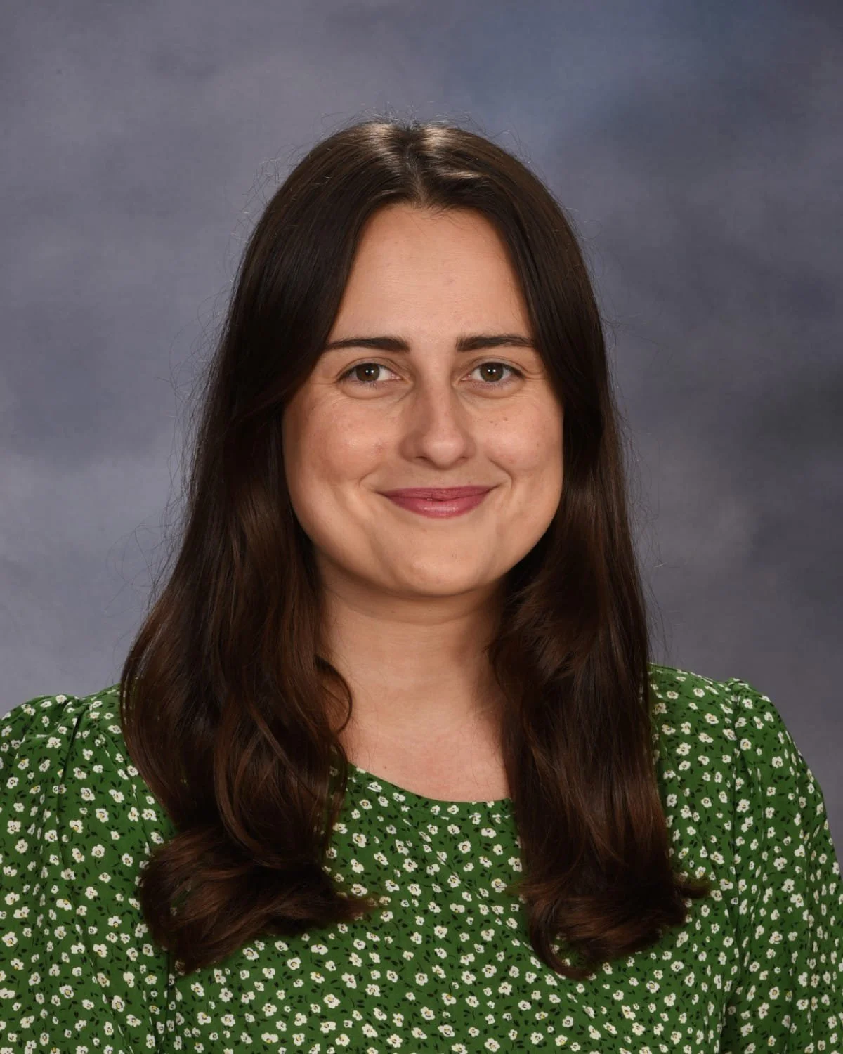 A woman with long dark hair wearing a green dress with white floral pattern, smiling against a grey background.