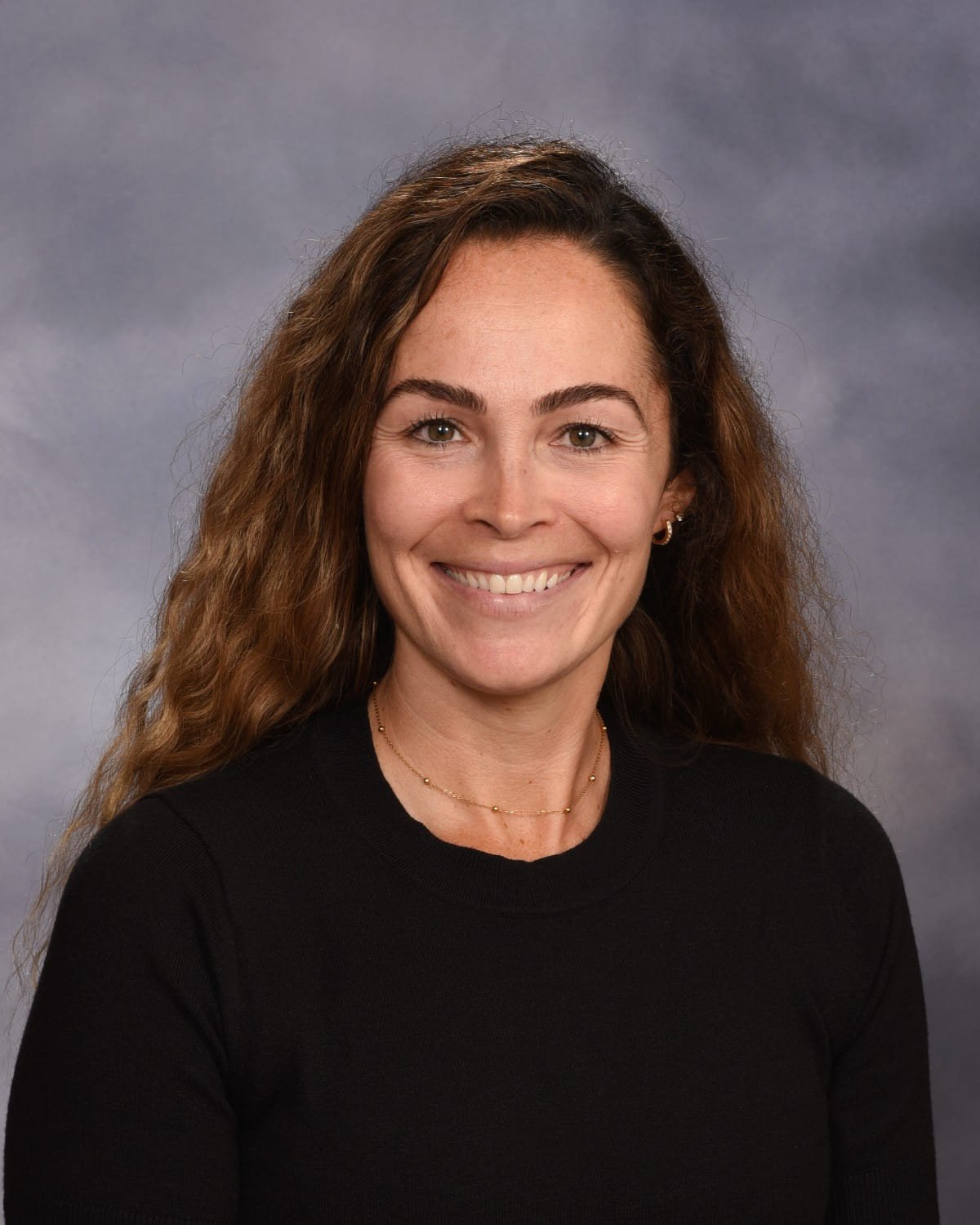 A woman with shoulder-length brown curly hair and a black top smiling at the camera against a gray background.