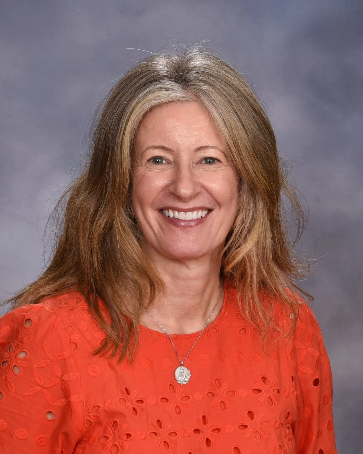 A middle-aged woman with long, light brown and blonde hair smiling at the camera, wearing a red blouse with eyelet floral patterns and a silver necklace, against a gray background.