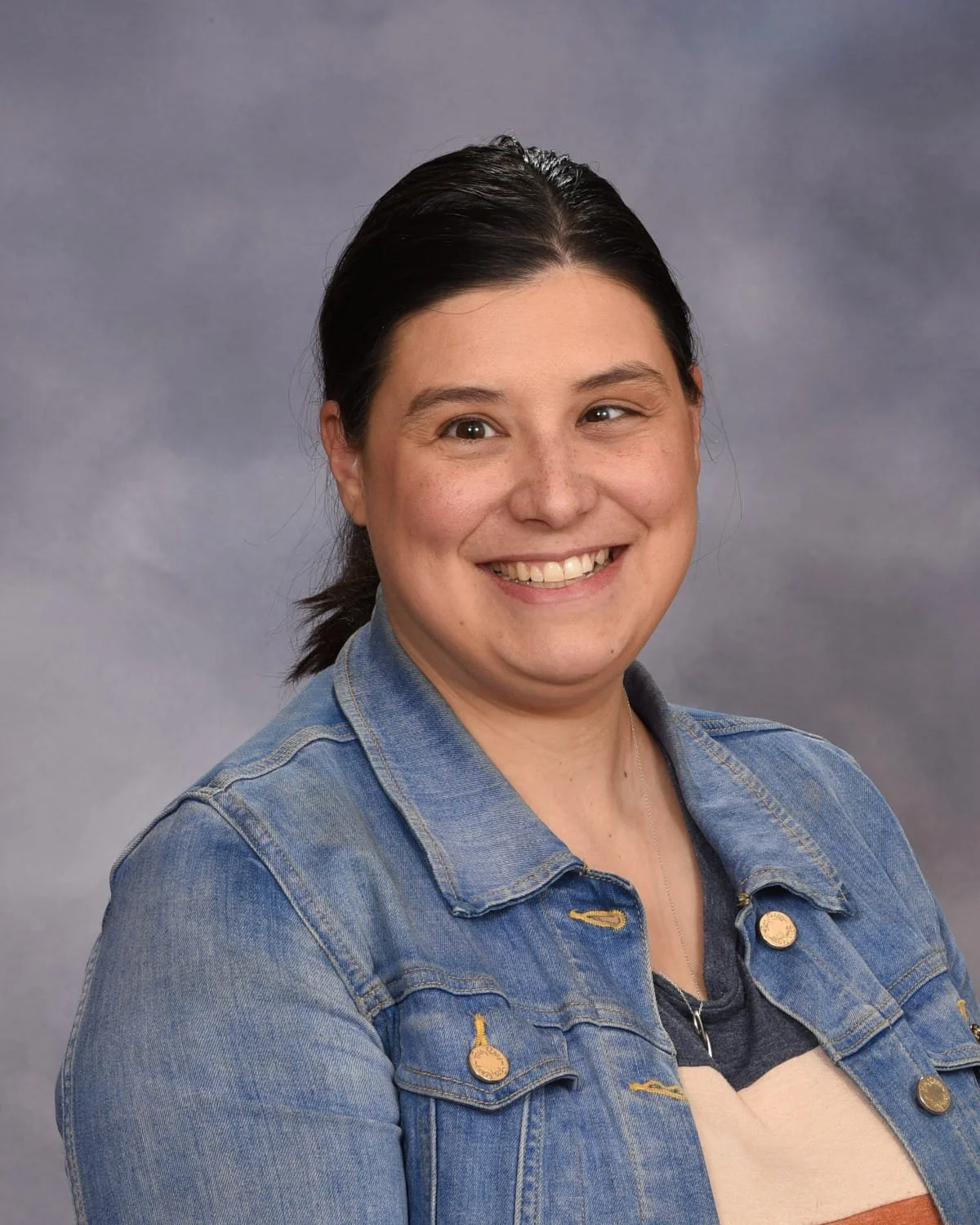 A woman with dark hair in a ponytail smiling at the camera, wearing a denim jacket over a striped shirt, against a gray background.