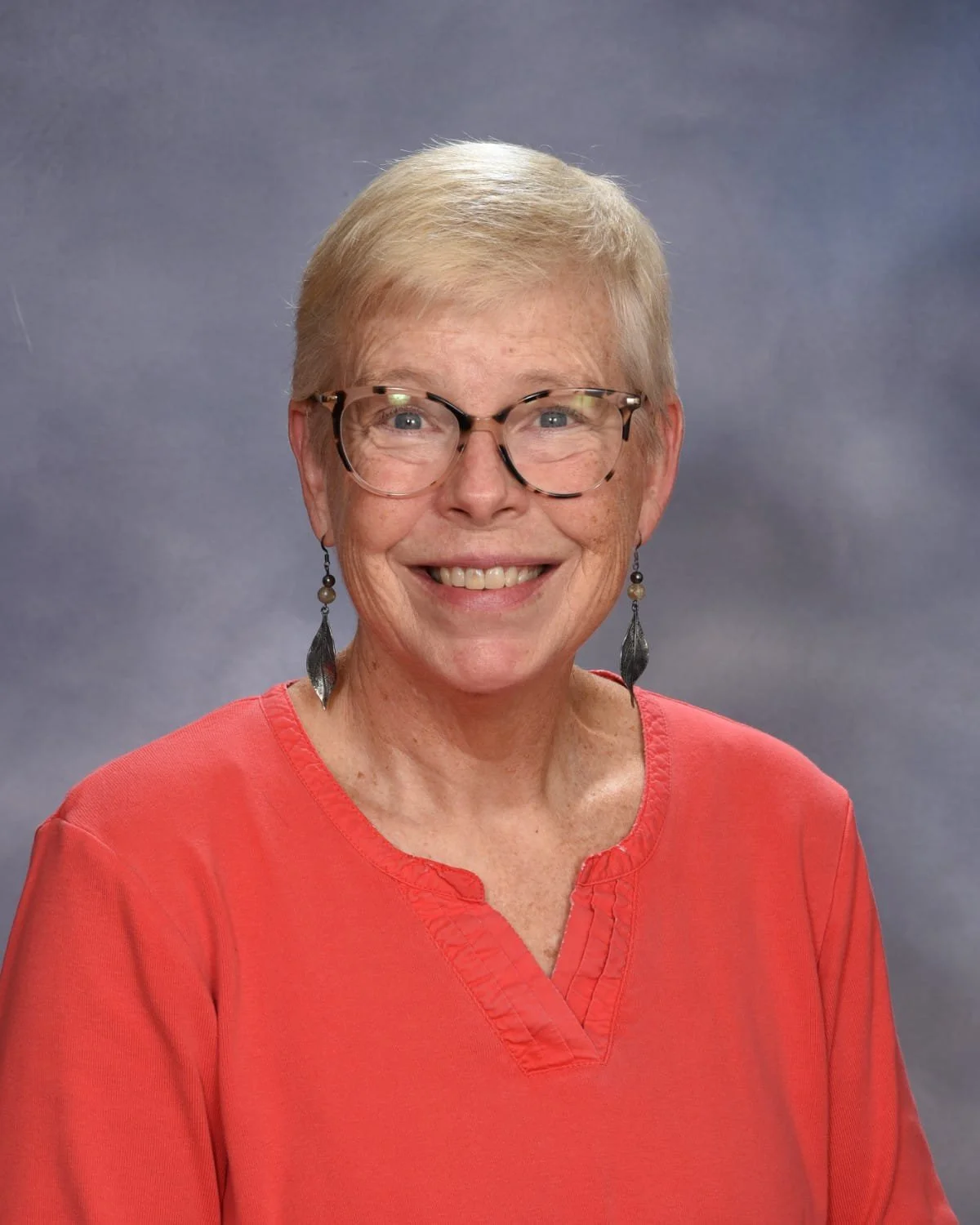 A smiling woman with short blonde hair, glasses, and earrings, wearing a coral-red top, against a gray background.