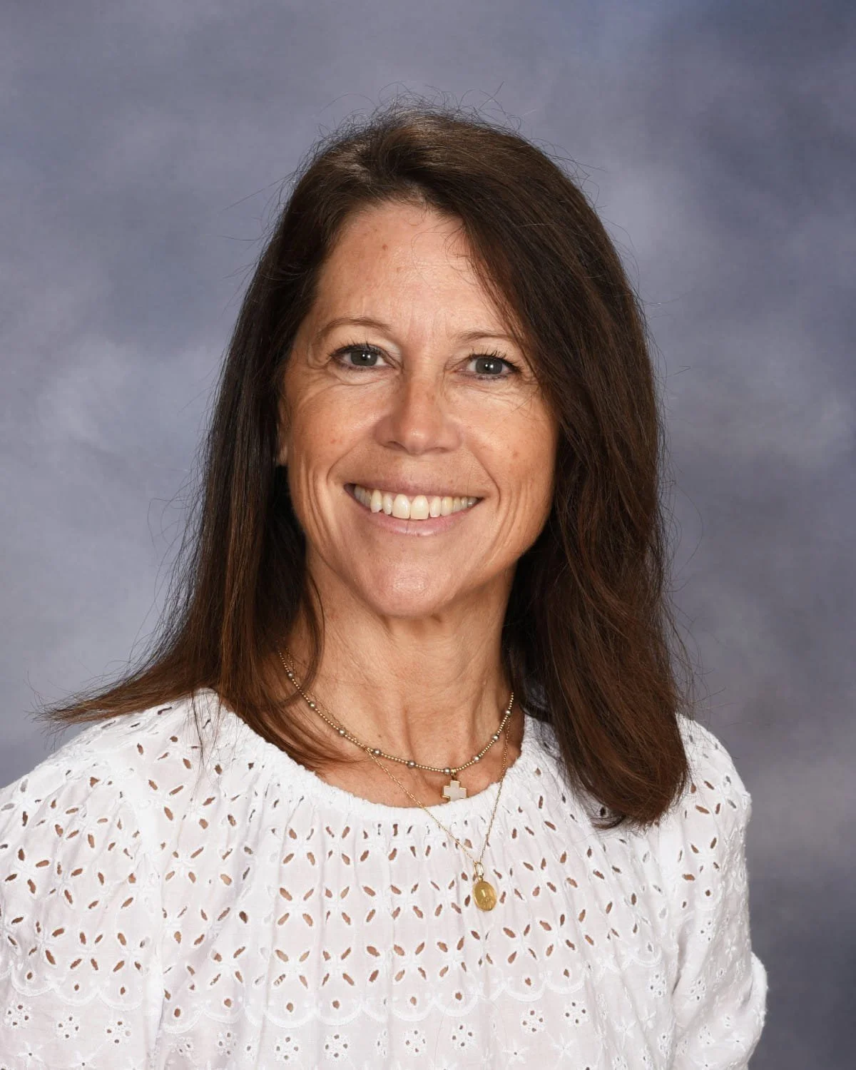 A woman with shoulder-length brown hair smiling, wearing a white eyelet blouse, two layered necklaces with cross and medallion pendants, against a gray background.