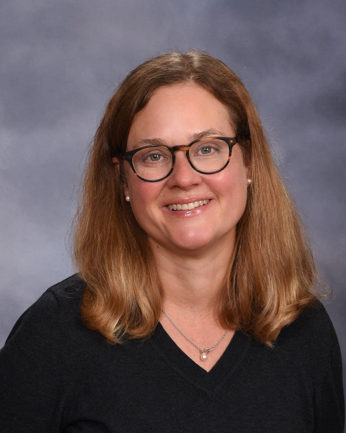 A woman with shoulder-length light brown hair, wearing black glasses, pearl earrings, a pearl necklace, and a black top, smiling in front of a gray background.