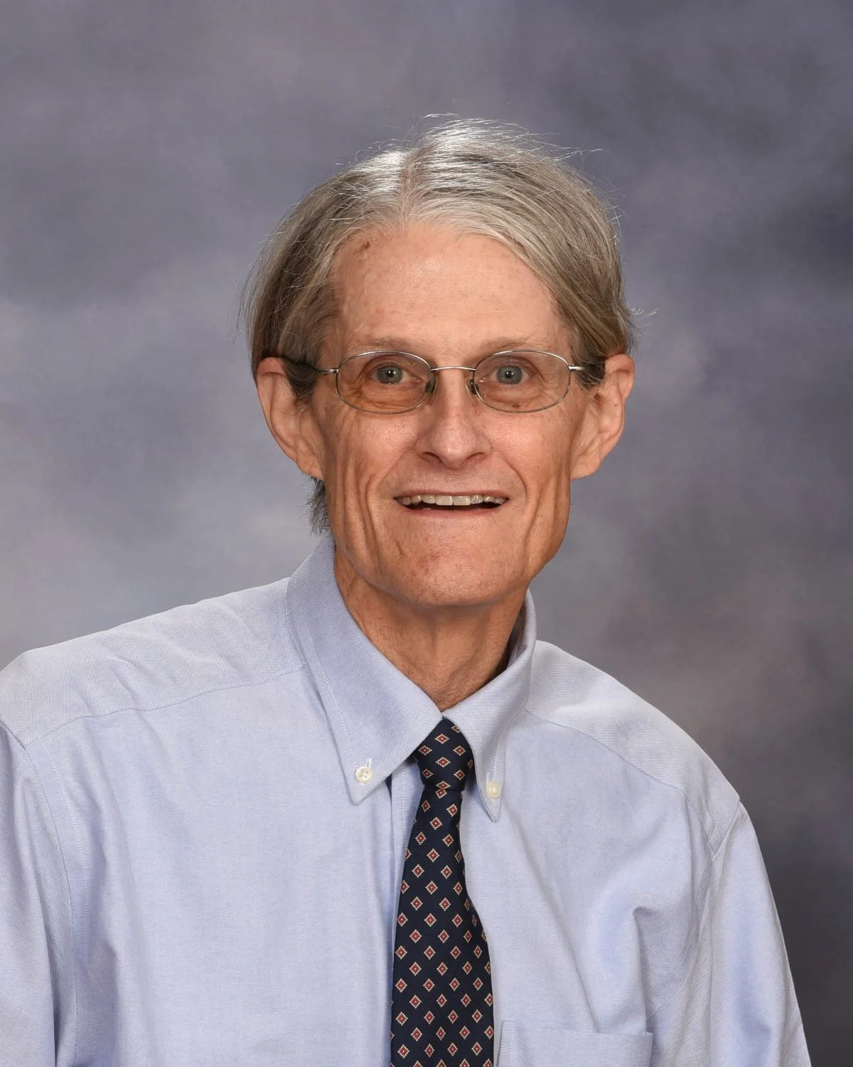 A middle-aged man with glasses, gray hair, wearing a light blue button-up shirt and a patterned tie, smiling against a neutral gray background.
