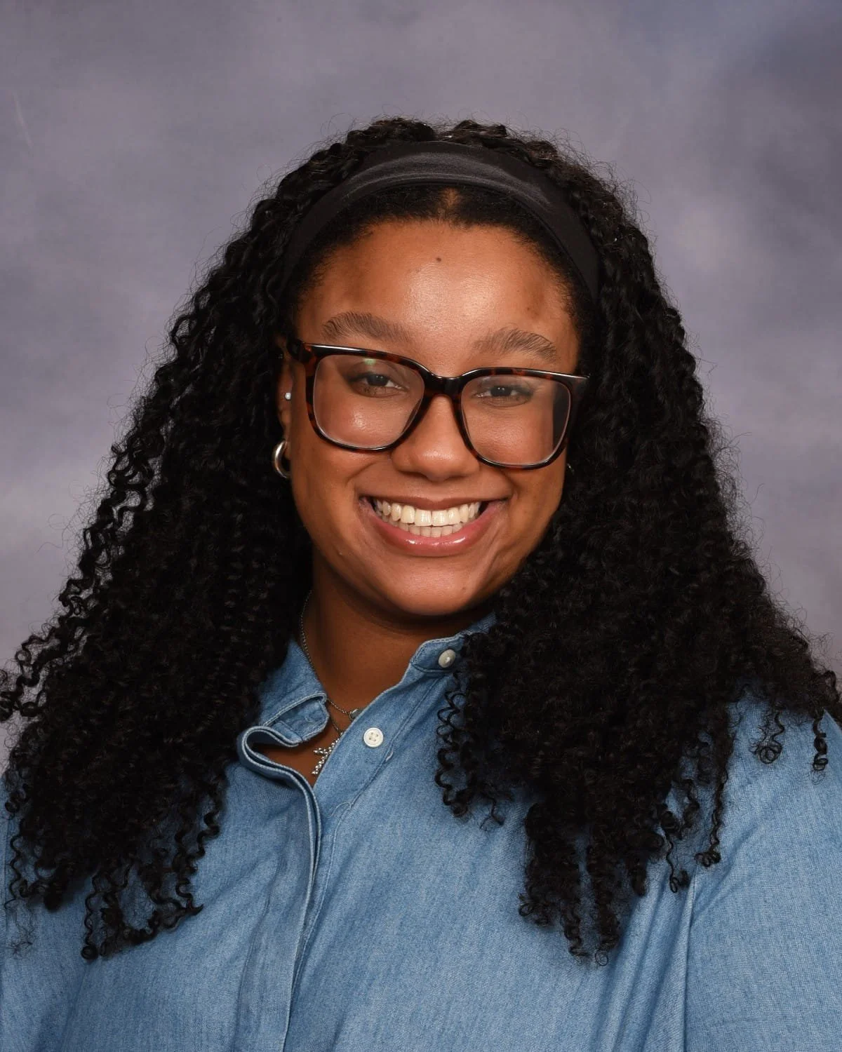 A woman with curly black hair, glasses, and a big smile, wearing a denim shirt against a gray backdrop.