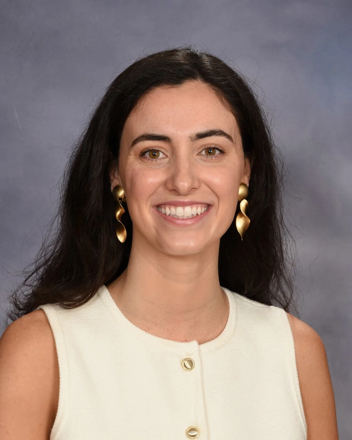 A woman with long dark hair wearing gold earrings and a sleeveless cream-colored top, smiling in front of a gray background.