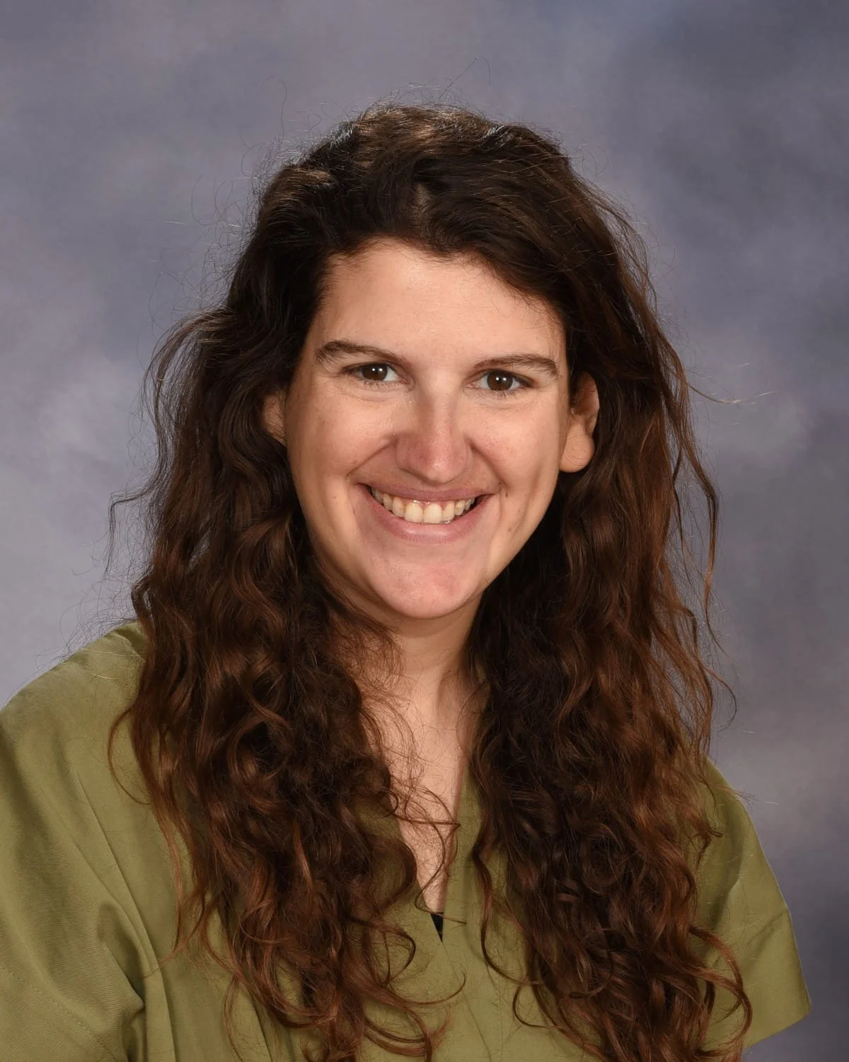 A woman with long, wavy brown hair and a bright smile, wearing a green top, against a gray background.
