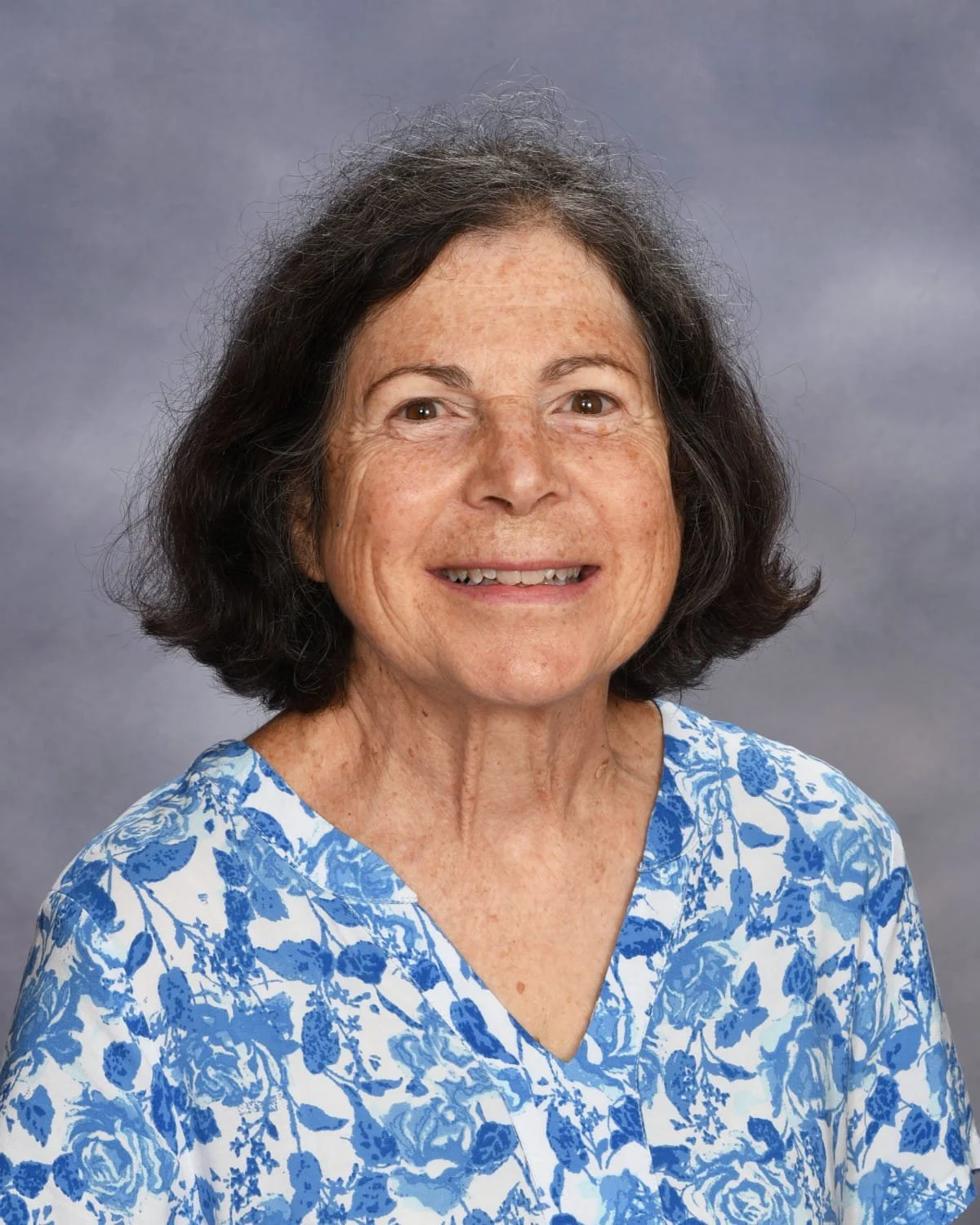 A smiling woman with shoulder-length dark hair and freckles, wearing a blue and white floral blouse, posed against a gray background.